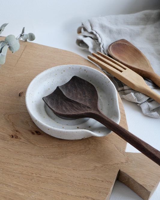 Wooden spoon on a white speckled dish on a wooden surface with a fork and cloth in the background.