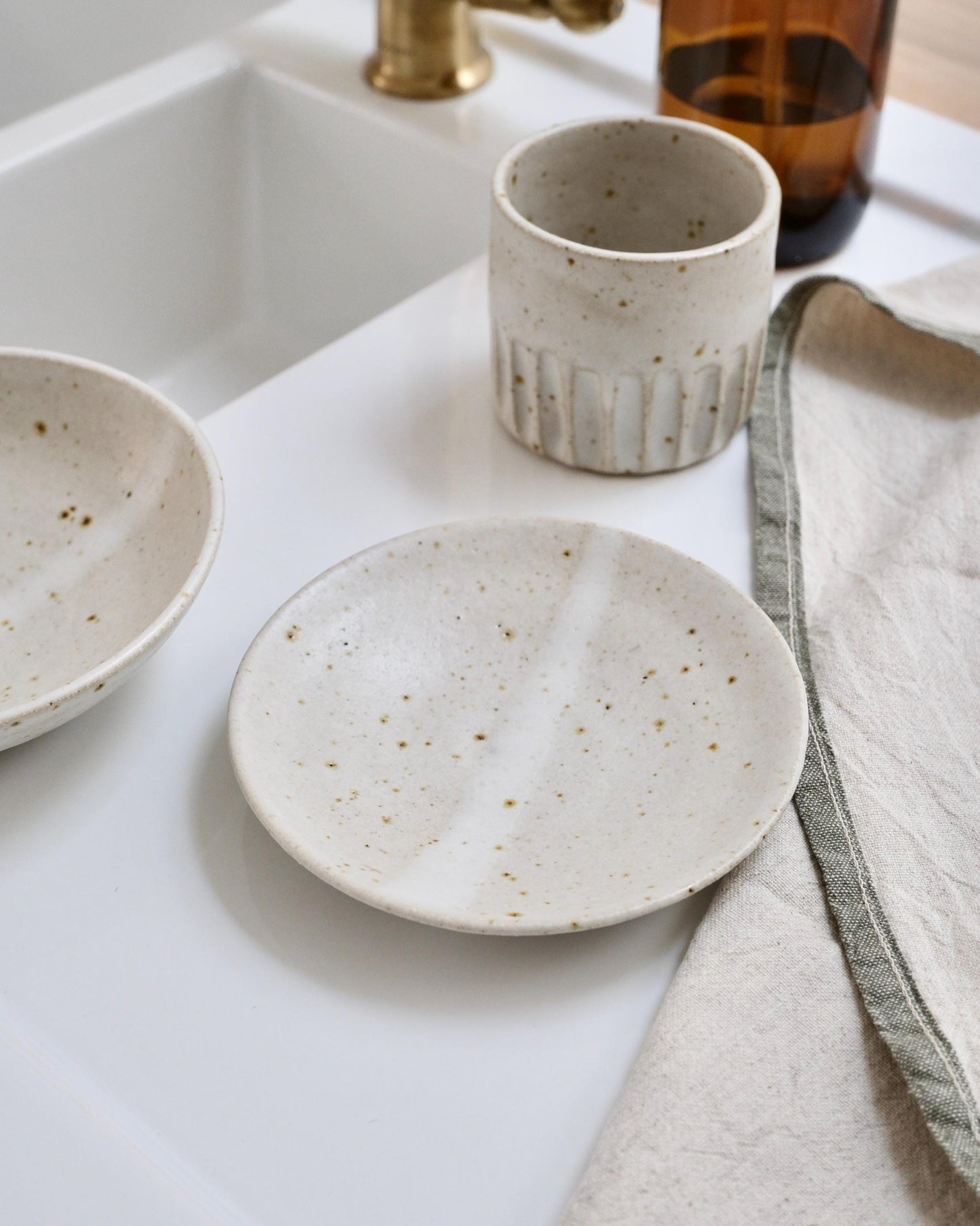 white Speckled ceramic dishes and cups on a white surface with a towel and bottle in the background.
