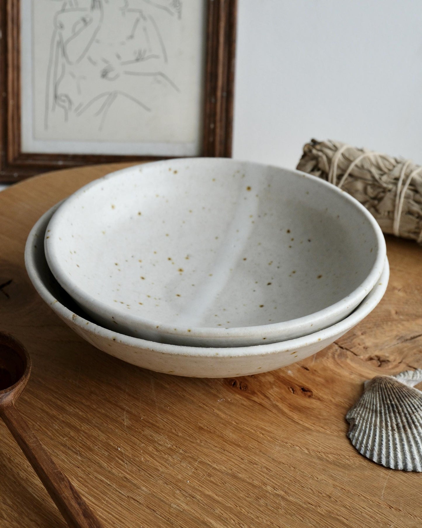 Two ceramic bowls stacked on a wooden surface with a framed picture and shell in the background.