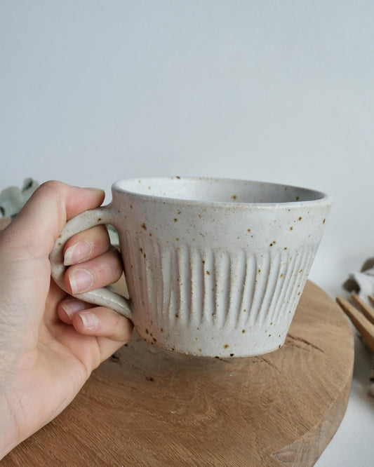 Hand holding a white speckled ceramic mug with a textured line surface pattern on a wooden surface.
