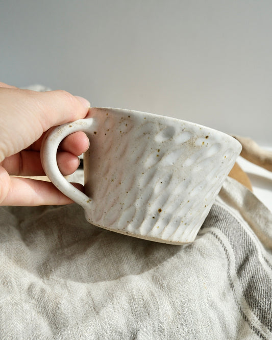 Hand holding a white textured ceramic coffee mug with a carved pattern on a tea towel