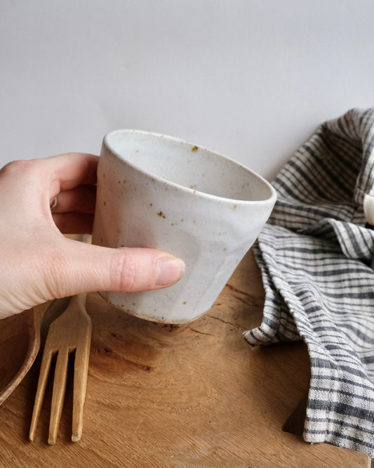 Hand holding a white ceramic cup on a wooden surface with a fork and checkered cloth in the background.