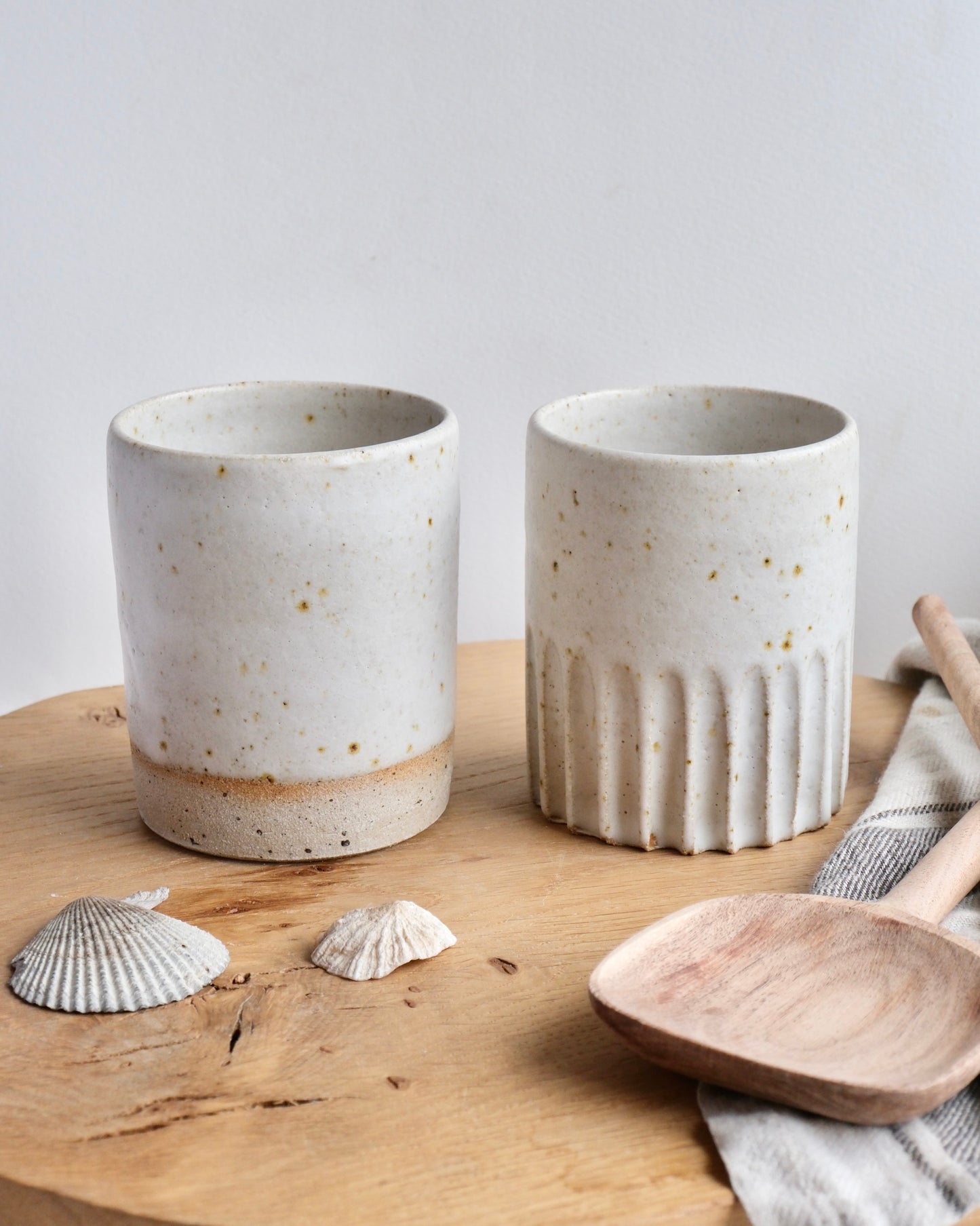 Two ceramic tall toothbrush holders on a wooden surface with shells and a wooden spoon.