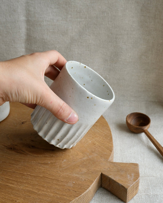 A hand holds a small ribbed ceramic tall pot with a speckled off-white glaze and gold flecks, set on a wooden board against a neutral linen background.