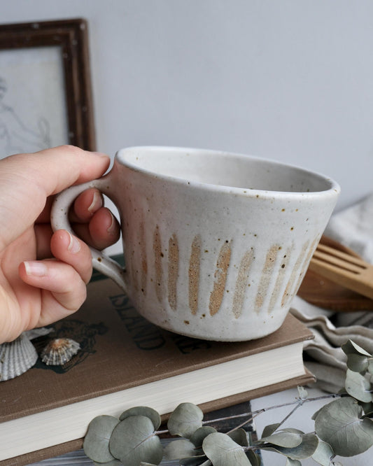 Hand holding a speckled striped ceramic mug with a textured surface on a table with books and decorative items.