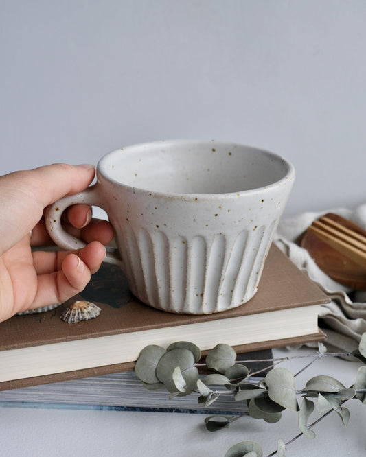 Hand holding a white ceramic tea mug on a surface with books and eucalyptus leaves.