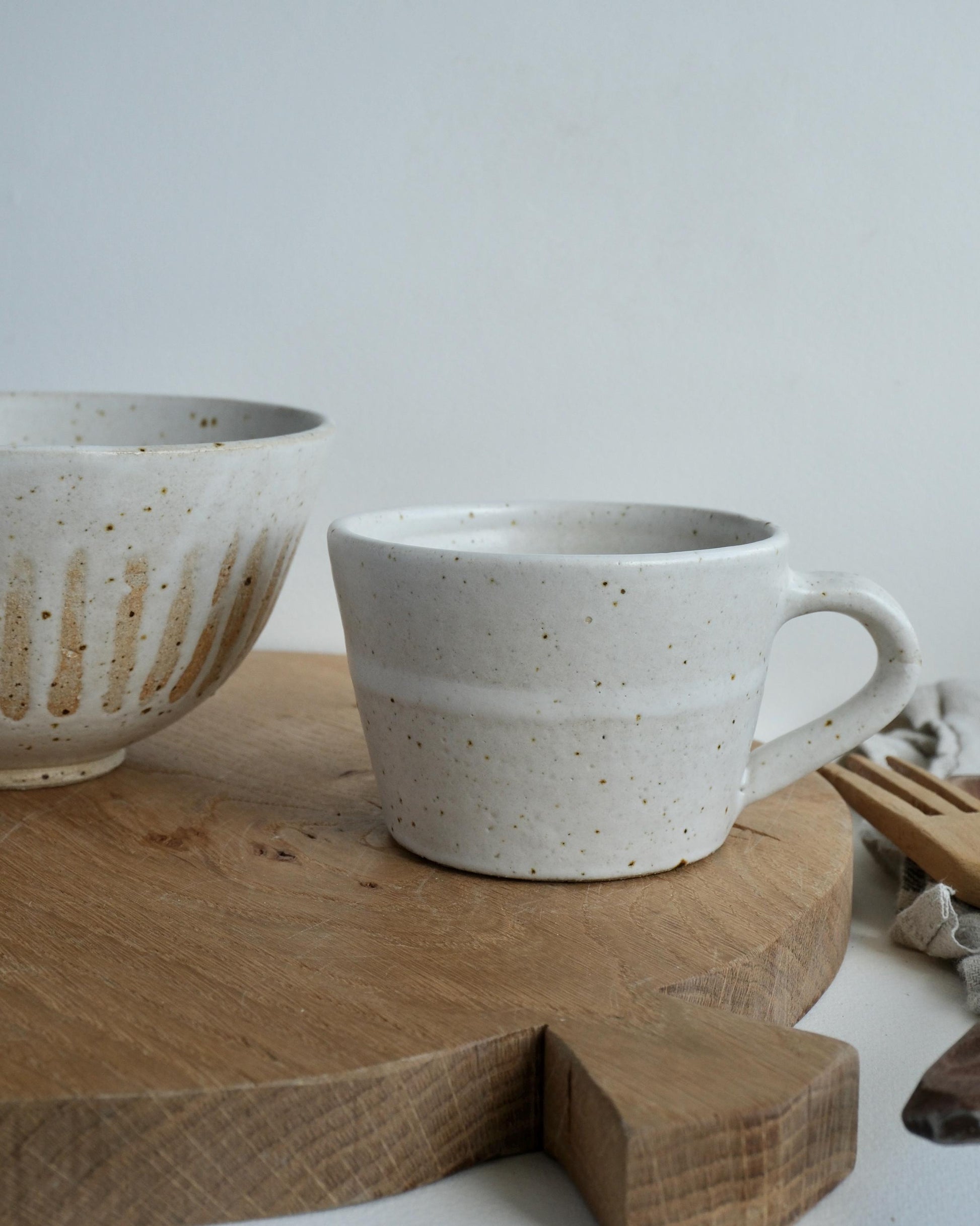 Speckled white ceramic mug and bowl on a wooden surface with a neutral background