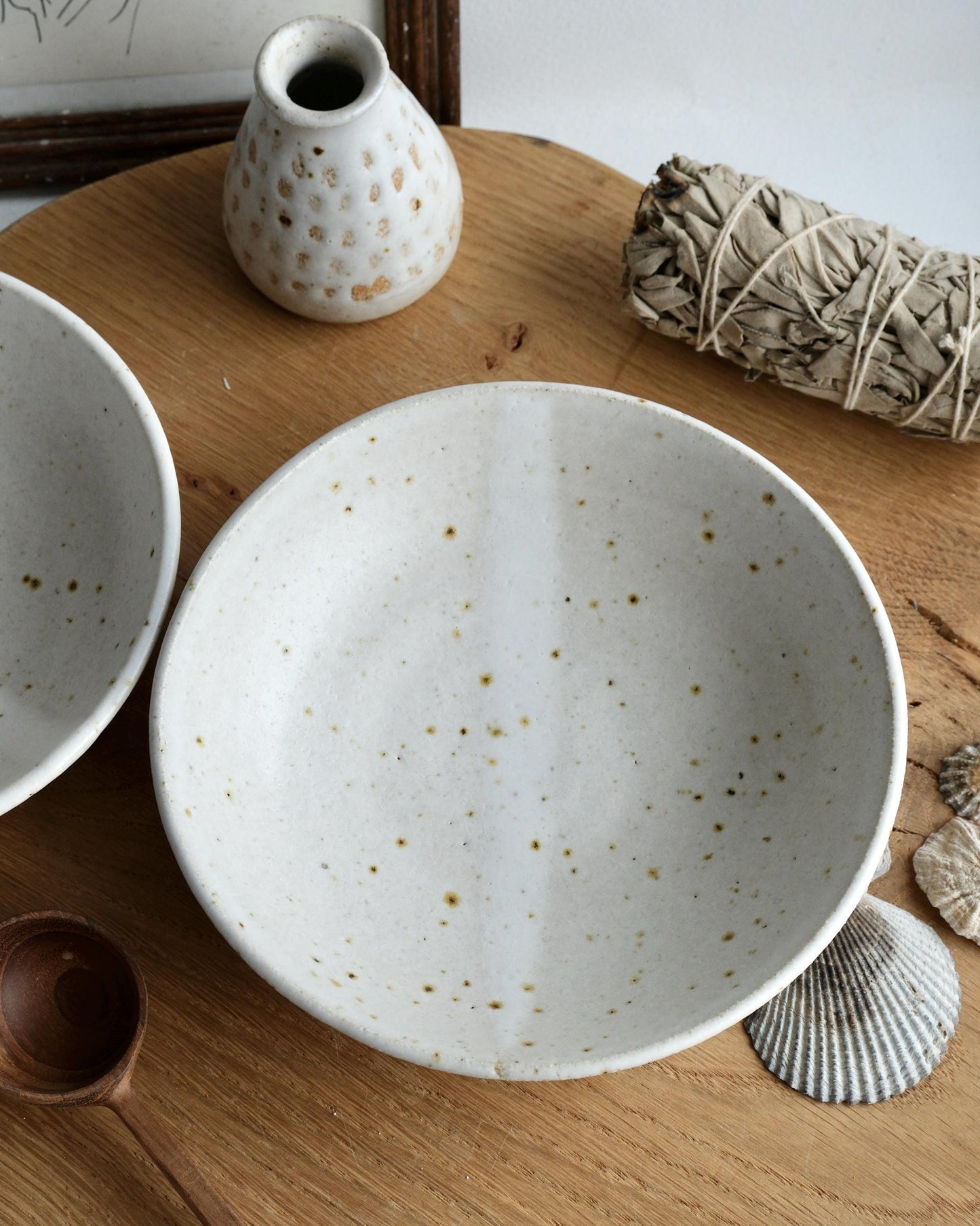 Speckled ceramic bowls on a wooden surface with a small vase and bundle of sage.