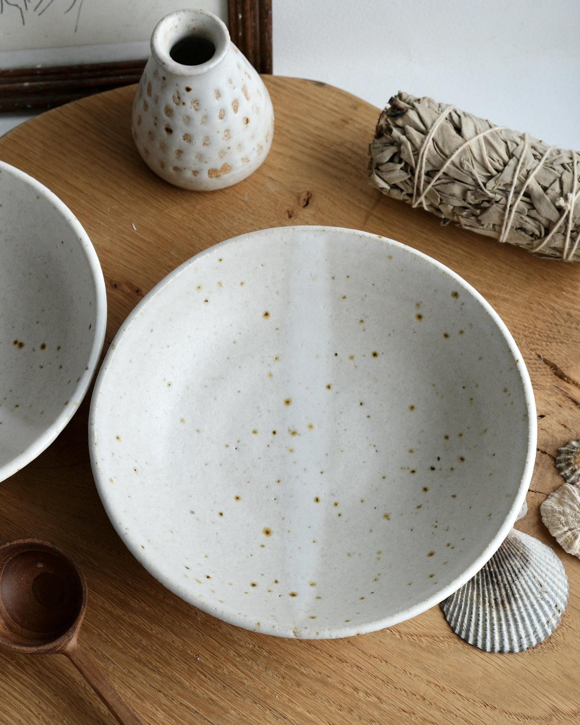 Speckled ceramic bowls on a wooden surface with a small vase and bundle of sage.