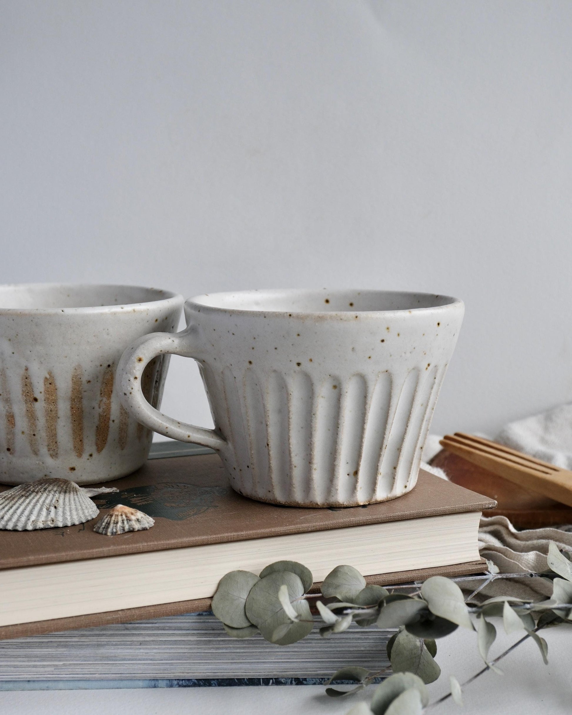 Two handmade ceramic mugs on a surface with books and decorative elements.