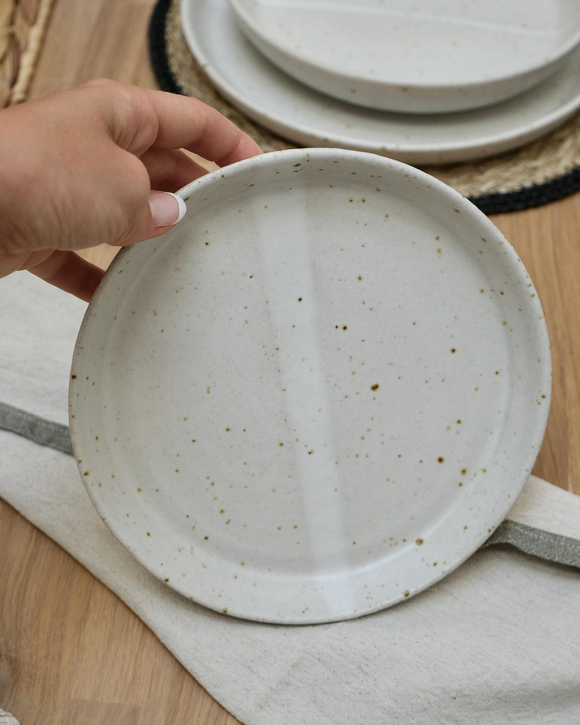 Hand holding a ceramic small side plate with speckled texture on a wooden surface