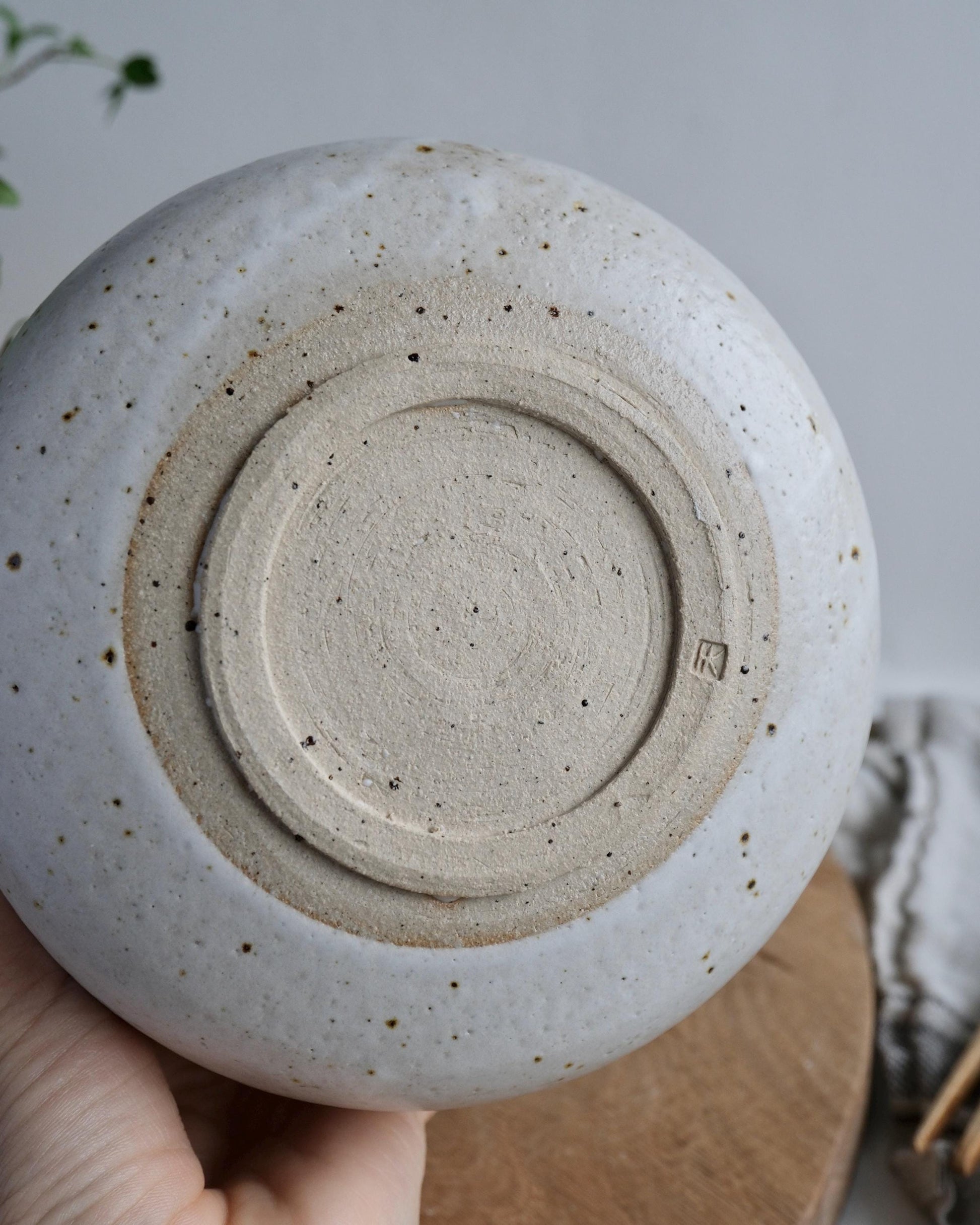 Close-up of the bottom of a white ceramic mixing bowl with a foot ring and makers mark, held by a hand against a neutral background.