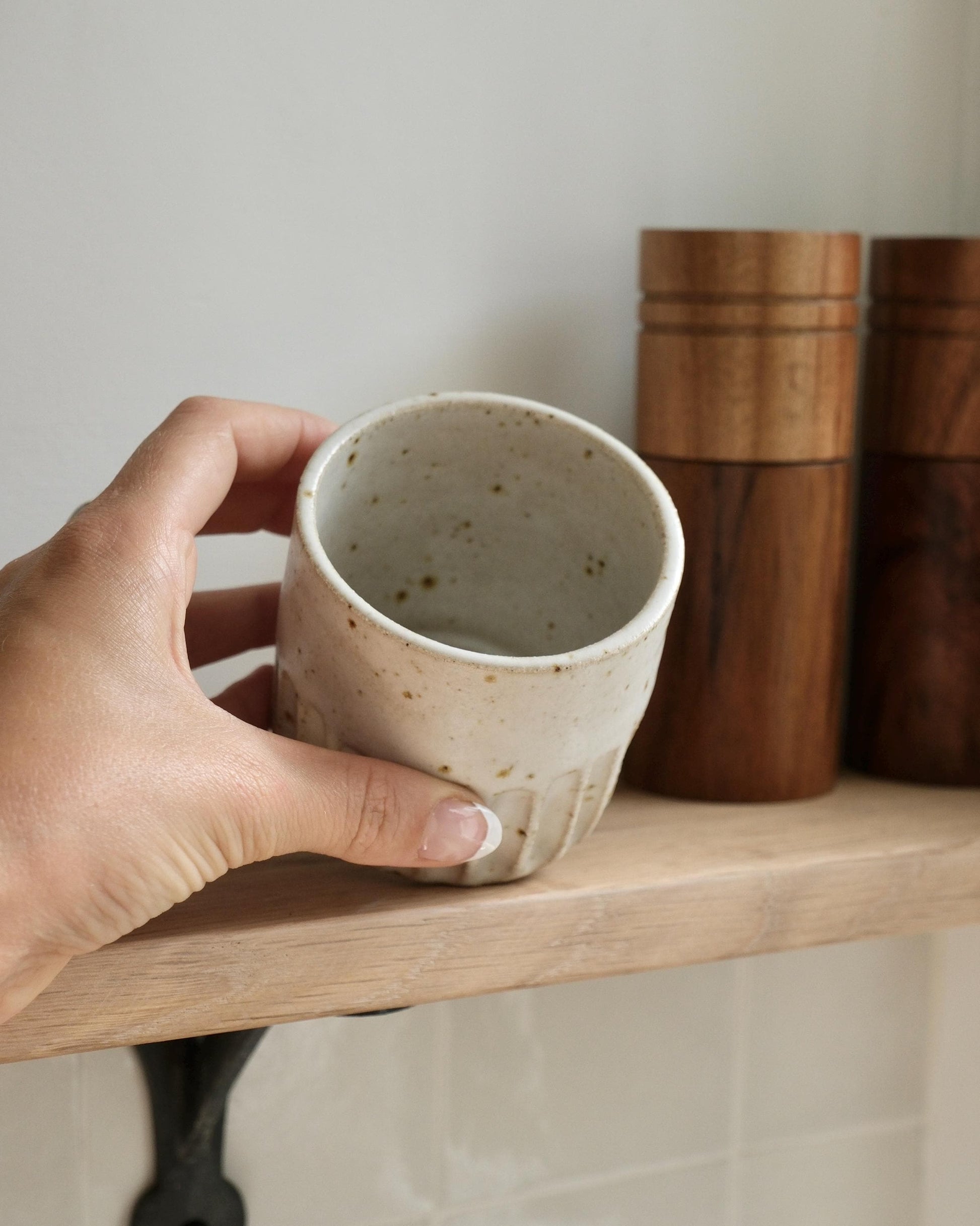 Hand holding a handmade white ceramic flat white cup on a wooden shelf with wooden containers in the background