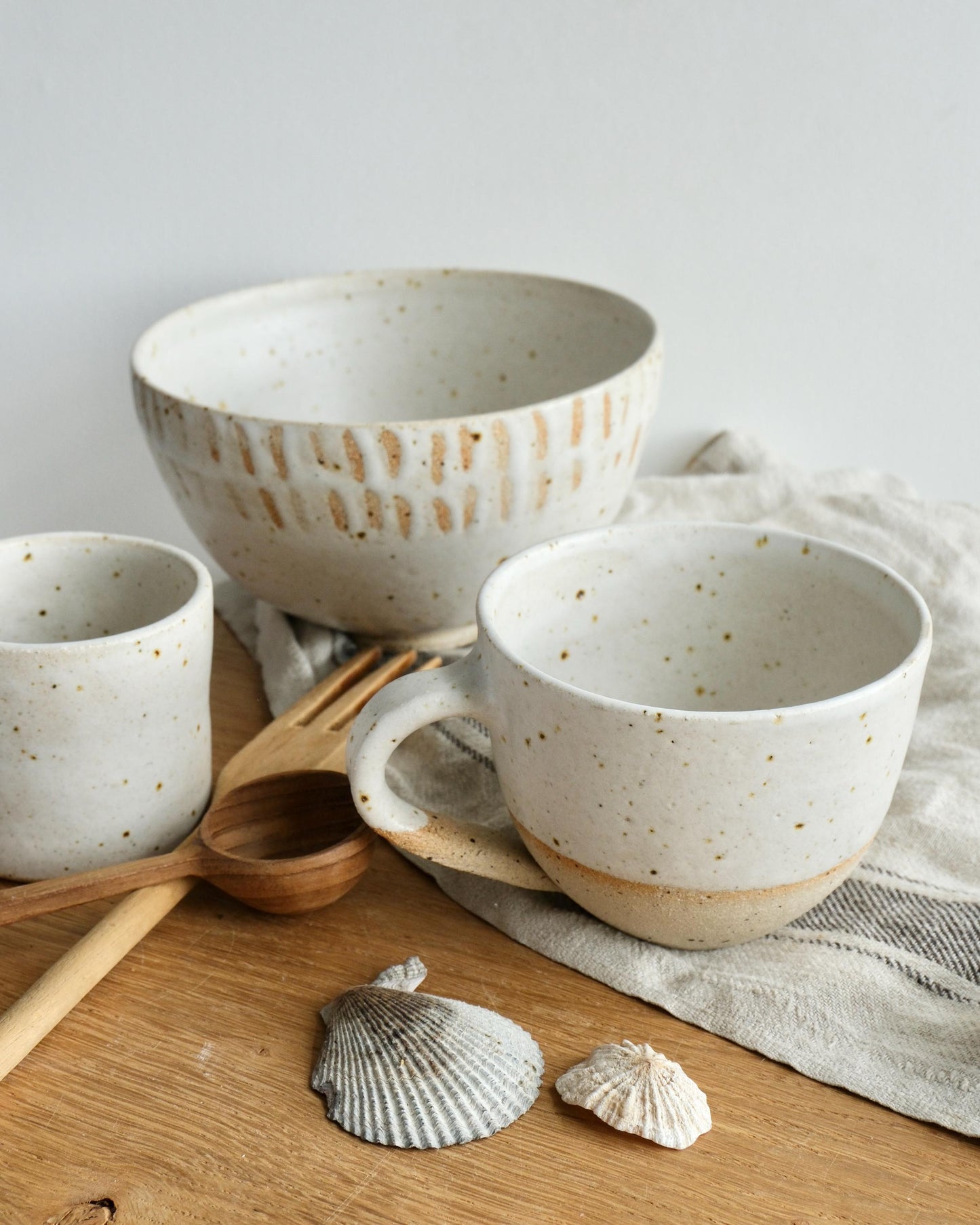 Set of ceramic bowls and mugs with white speckled pattern on a wooden surface with shells and a wooden spoon.