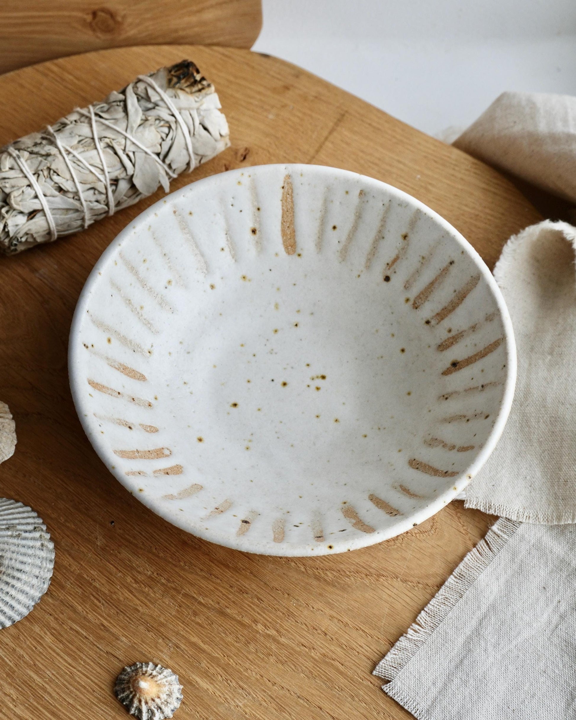 White speckled ceramic trinket bowl with an orange line pattern  design on a wooden surface with a white cloth and natural elements.