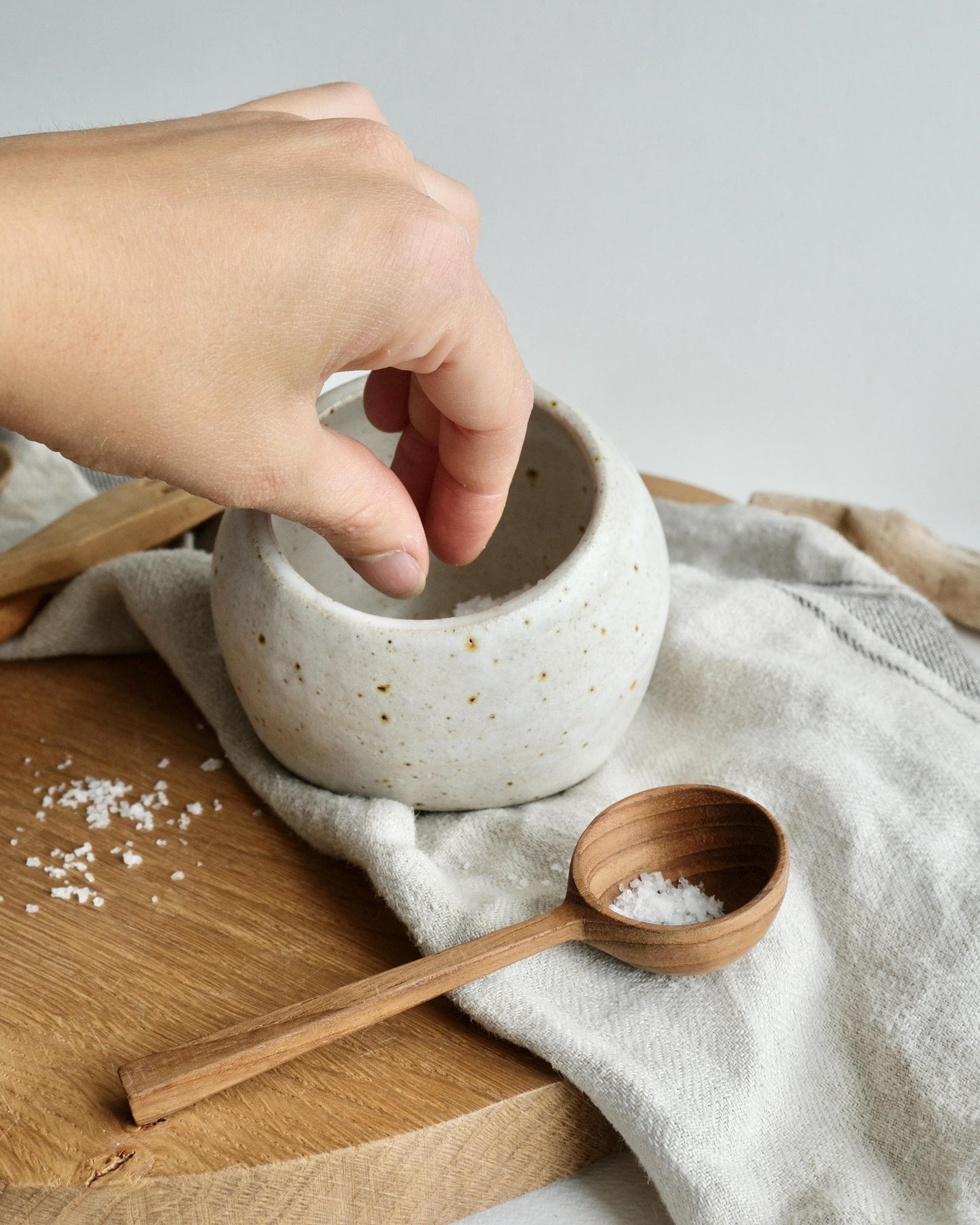 Hand reaching into a small white ceramic salt cellar bowl on a wooden surface with a wooden spoon nearby.