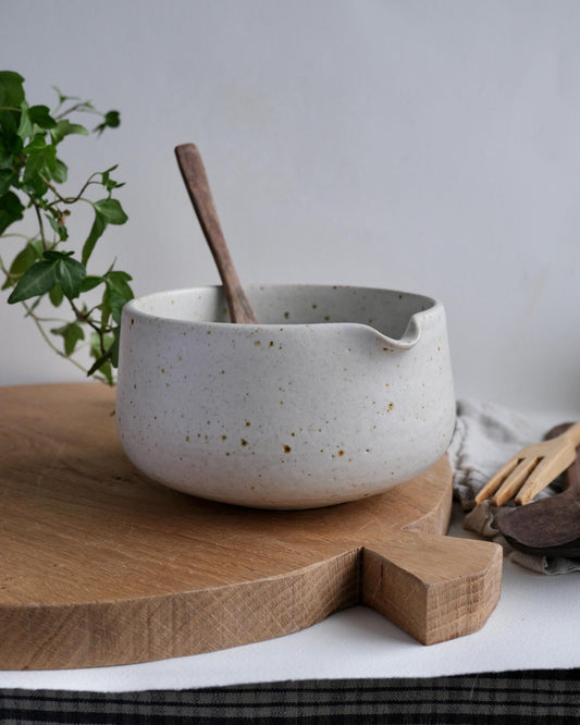 White ceramic matcha mixing bowl with speckled texture on a wooden cutting board, with a plant in the background.