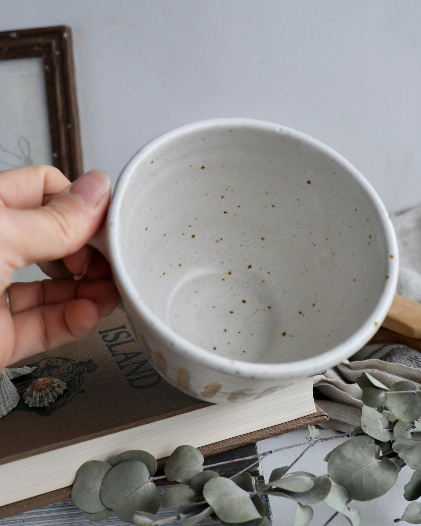 Hand holding a white ceramic mug with speckled pattern on a surface with books and plants.