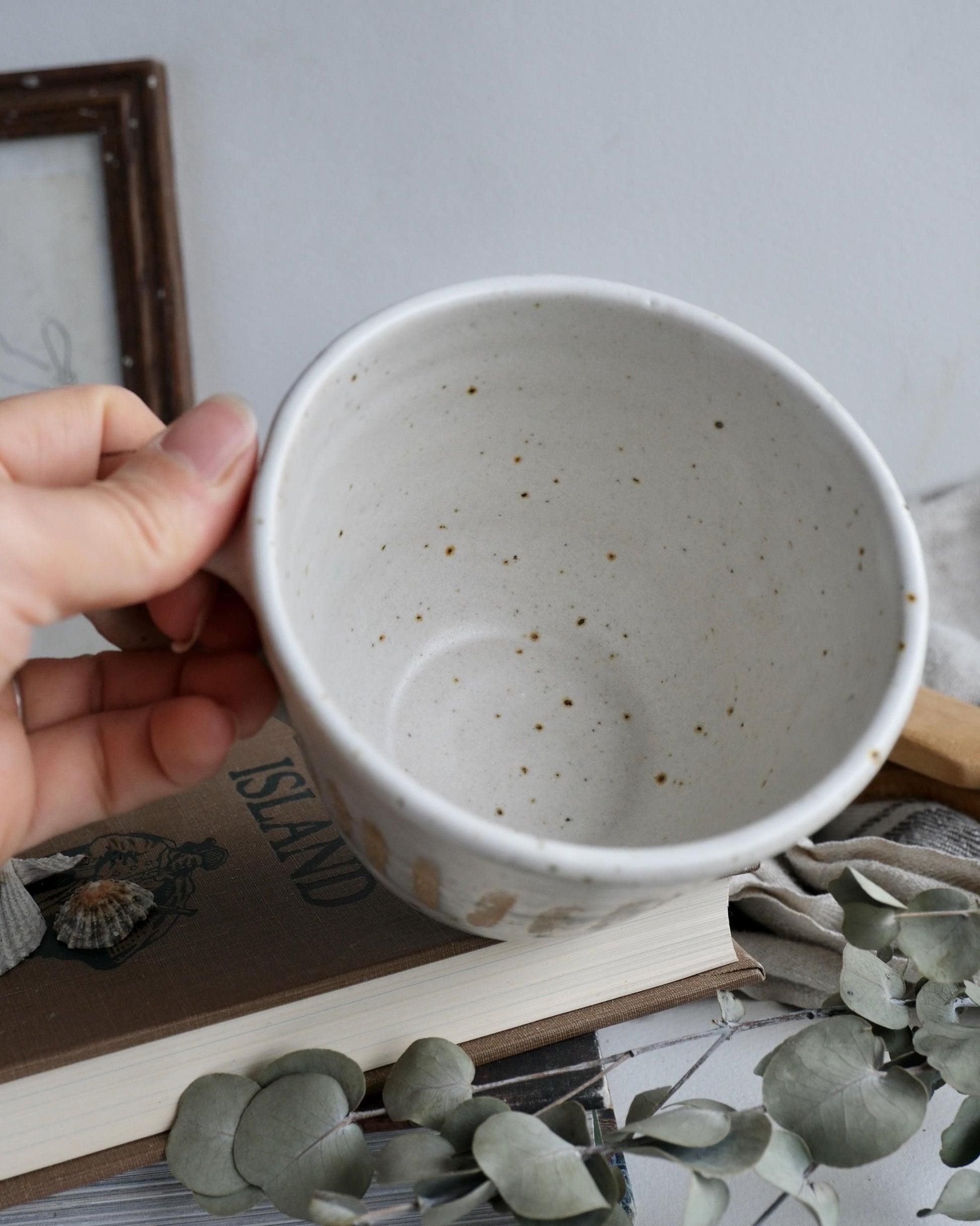 Hand holding a white ceramic mug with speckled pattern on a surface with books and plants.