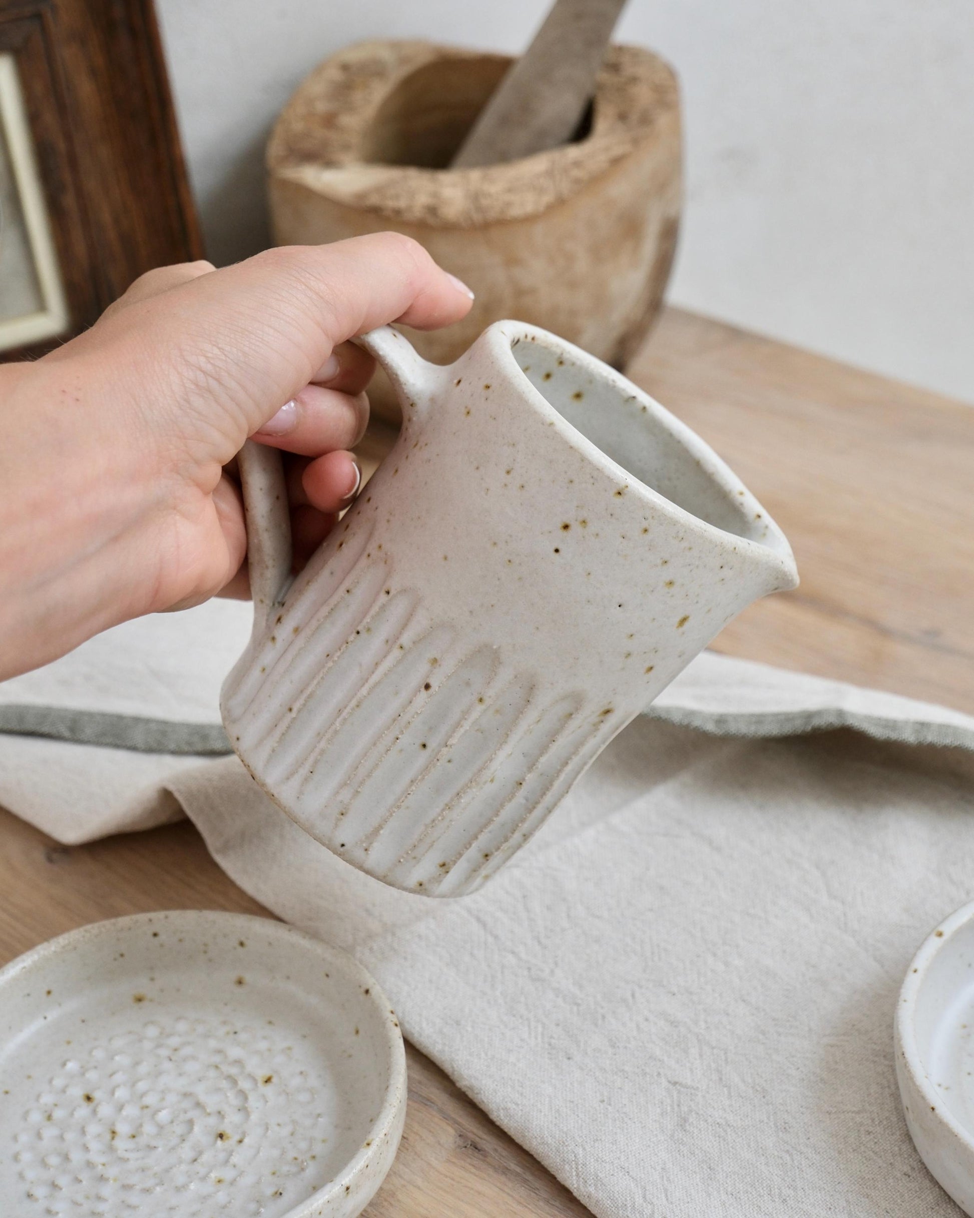 Person holding a white speckled ceramic milk jug with a line textured surface on a wooden table.
