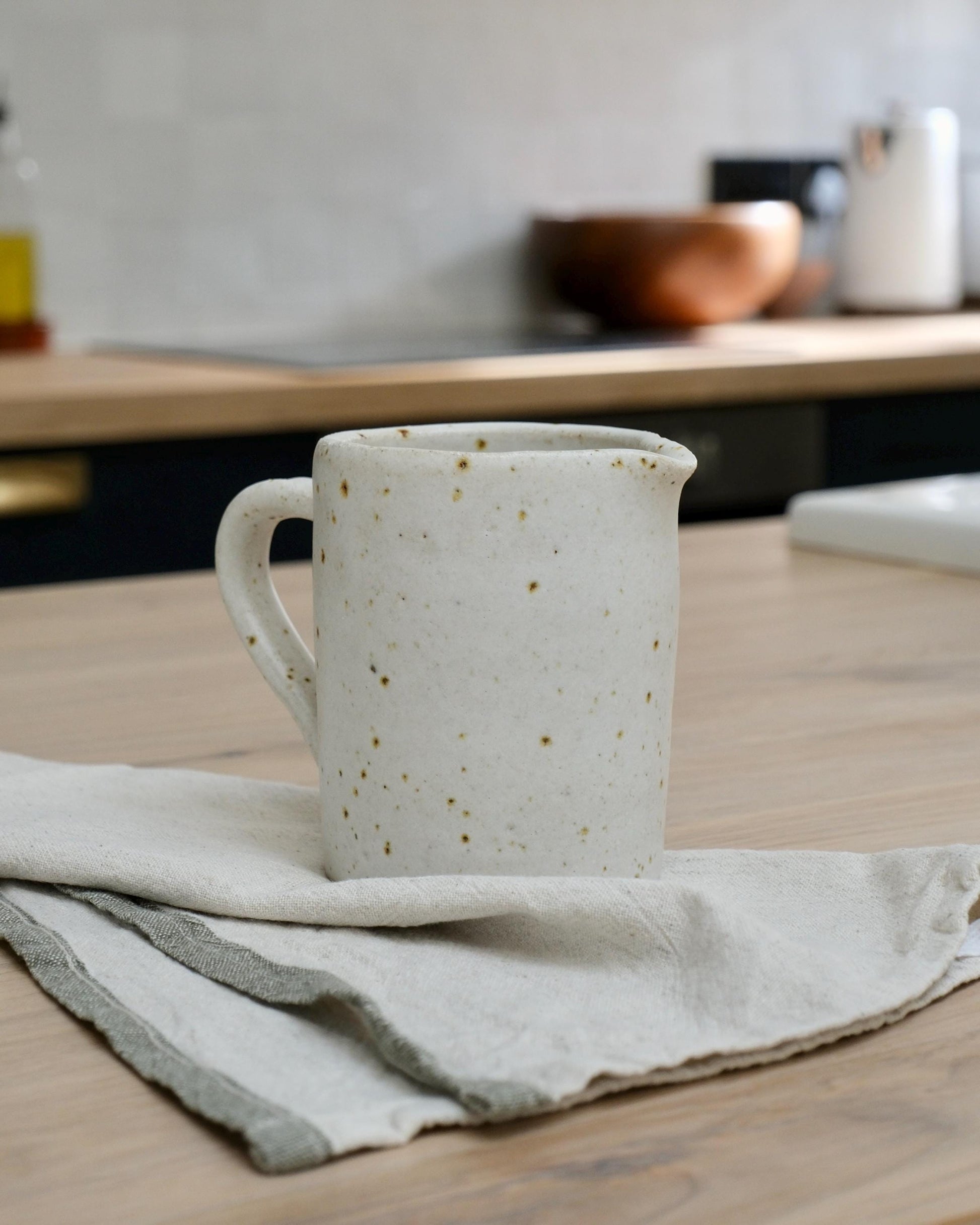 White speckled ceramic creamer on a wooden surface with a kitchen background