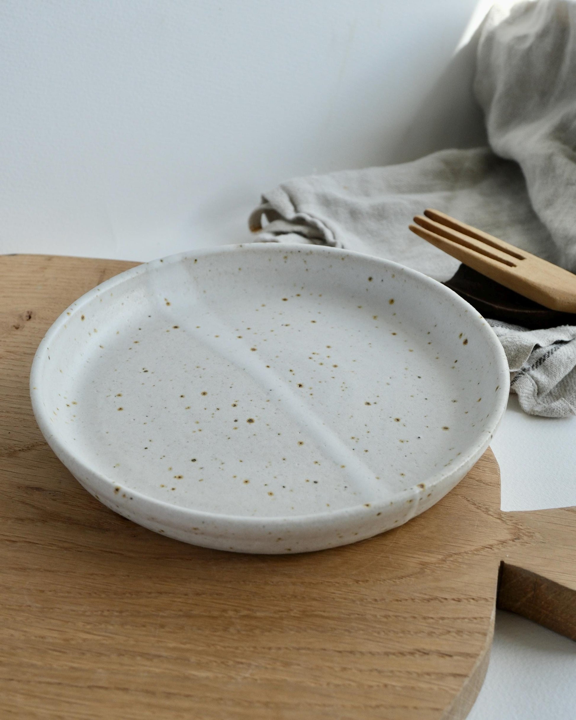 Handmade Speckled ceramic small side plate on a wooden surface with a cloth and cutlery in the background.