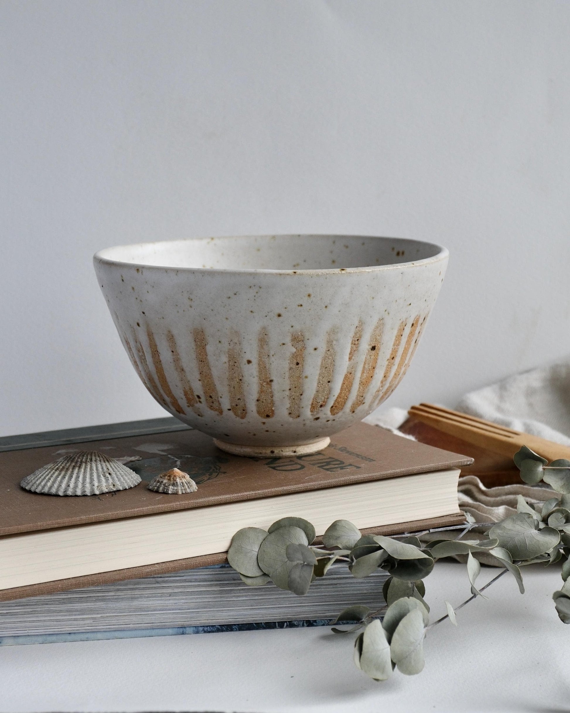 white Ceramic bowl with orange stripes on books with eucalyptus leaves and seashells on a light background