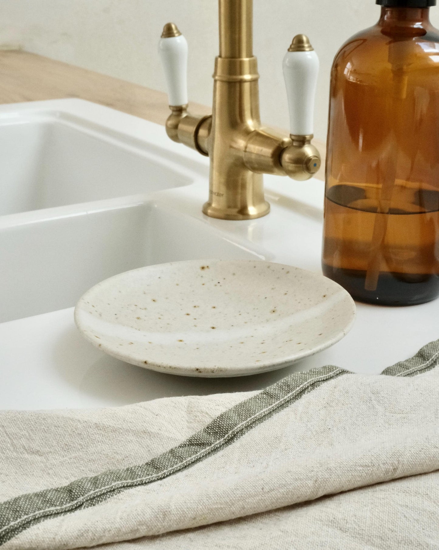 Kitchen sink with gold faucet, white ceramic soap dish, and brown bottle on a white countertop.
