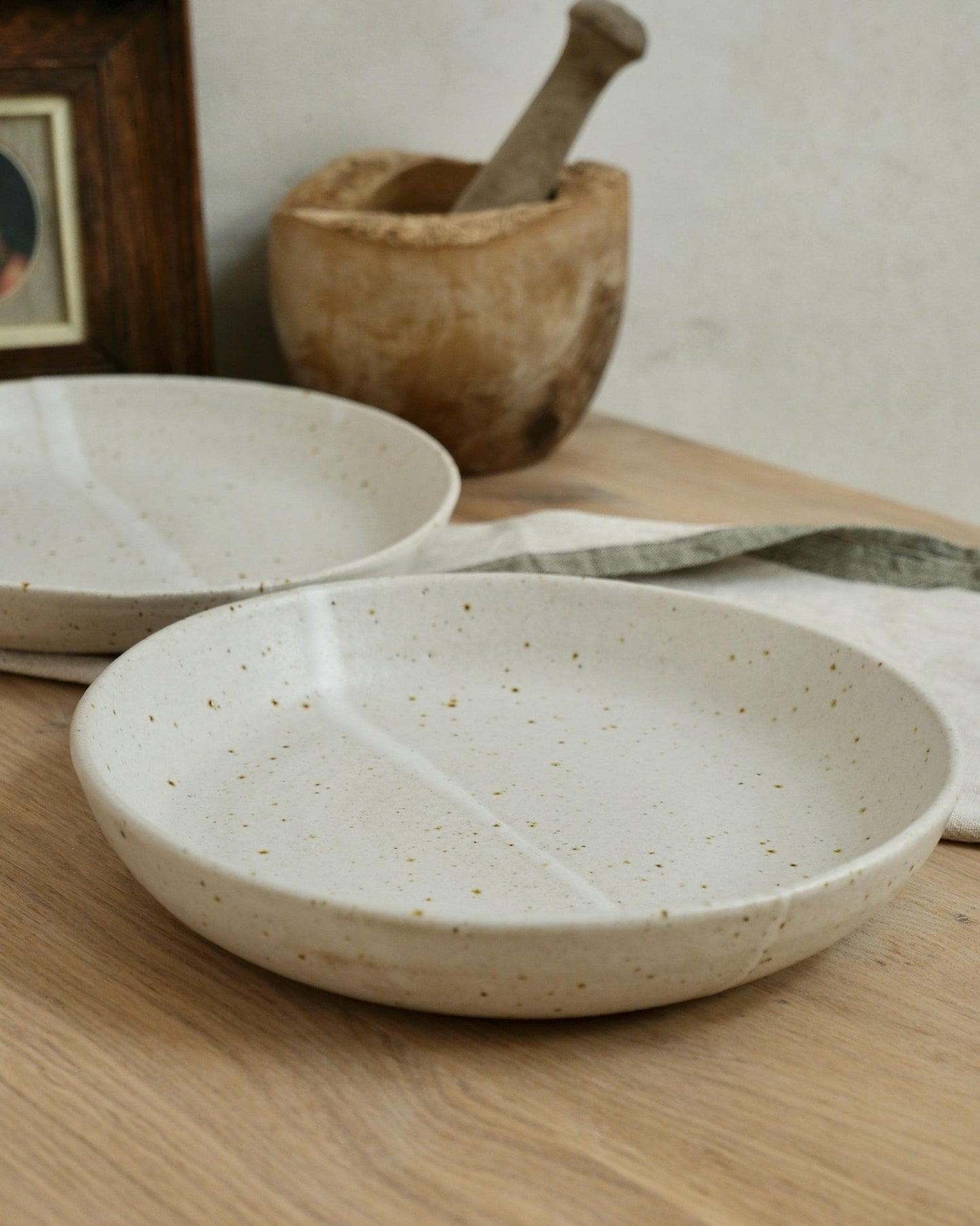 Two white ceramic pasta bowls on a wooden surface with a wooden mortar and pestle in the background.