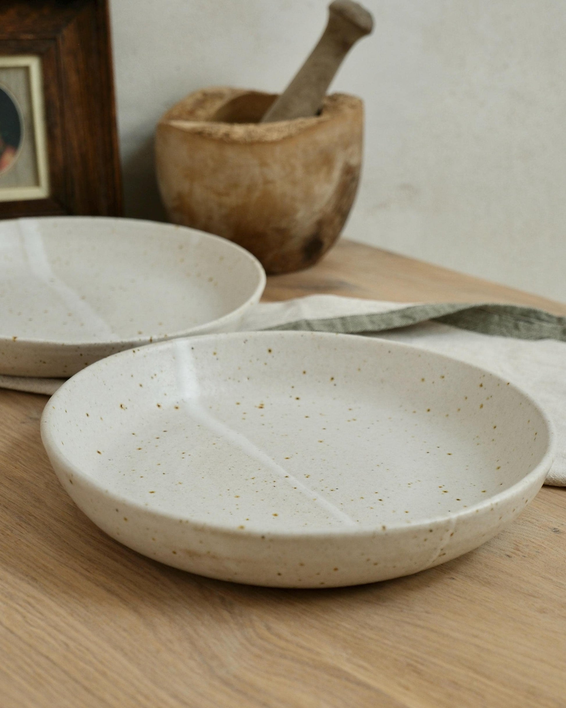 Two white ceramic pasta bowls on a wooden surface with a wooden mortar and pestle in the background.