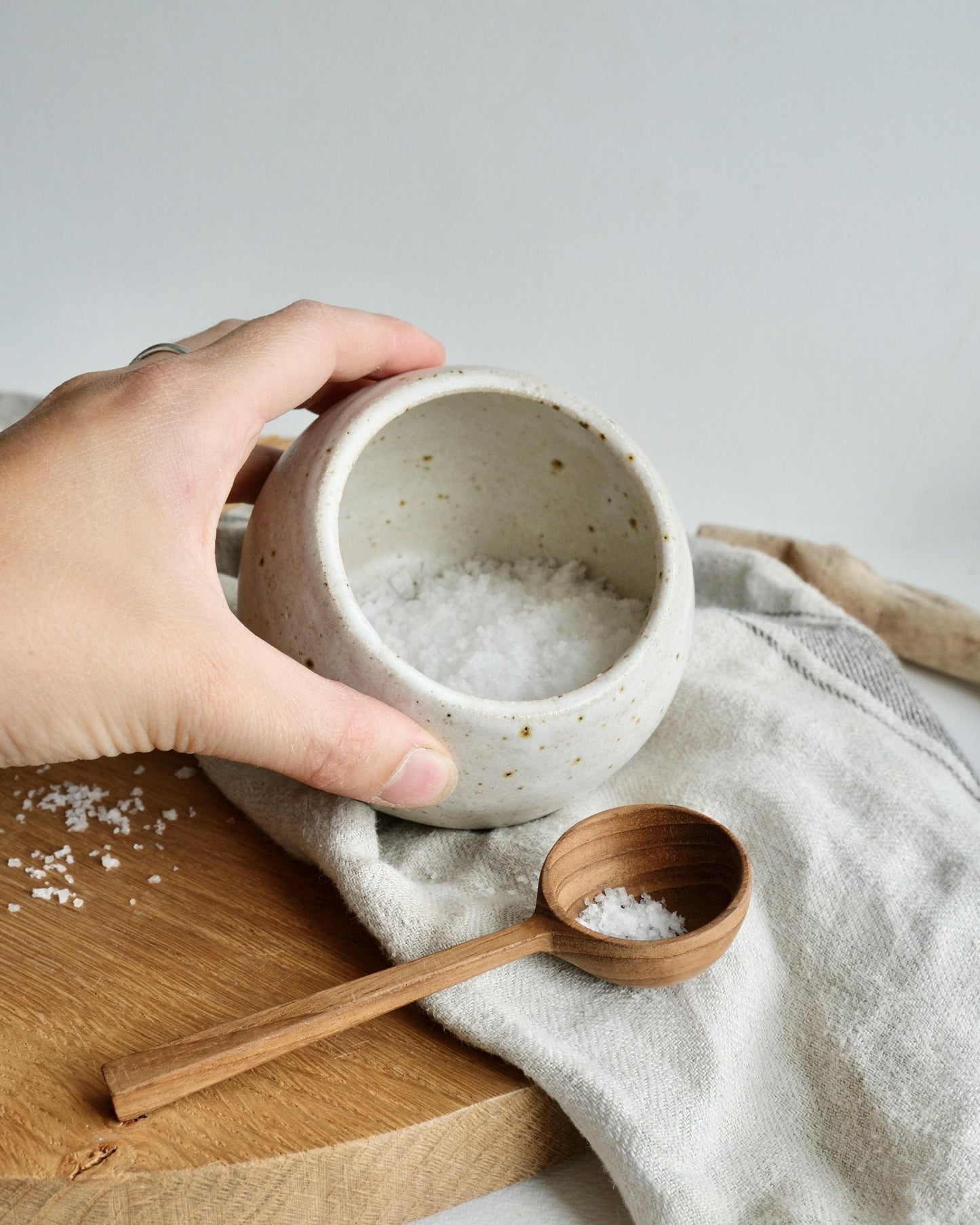 Hand holding a white ceramic salt cellar  bowl filled with salt, next to a wooden spoon on a wooden board and cloth.
