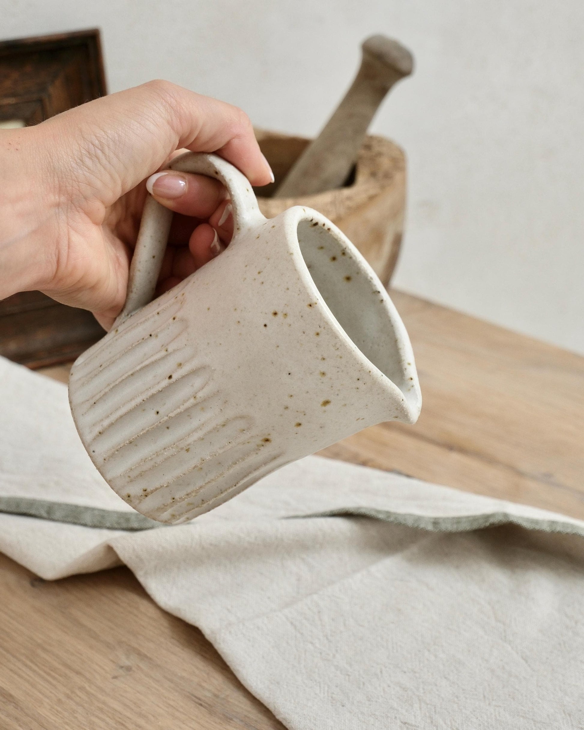 Hand holding a white speckled ceramic mug with a carved line pattern on a wooden surface with a neutral background
