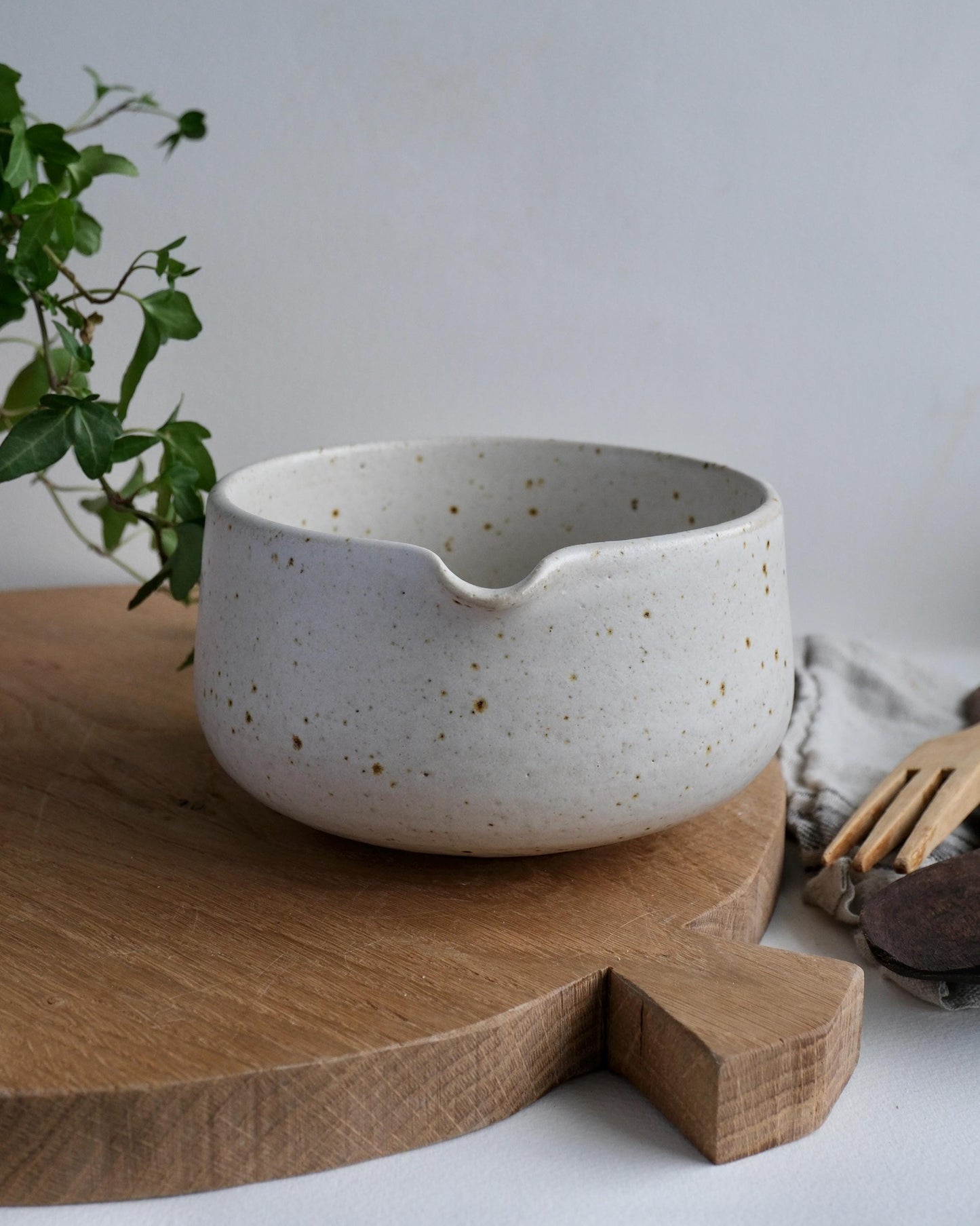 white Speckled ceramic mixing bowl on a wooden cutting board with a plant and kitchen towel in the background.