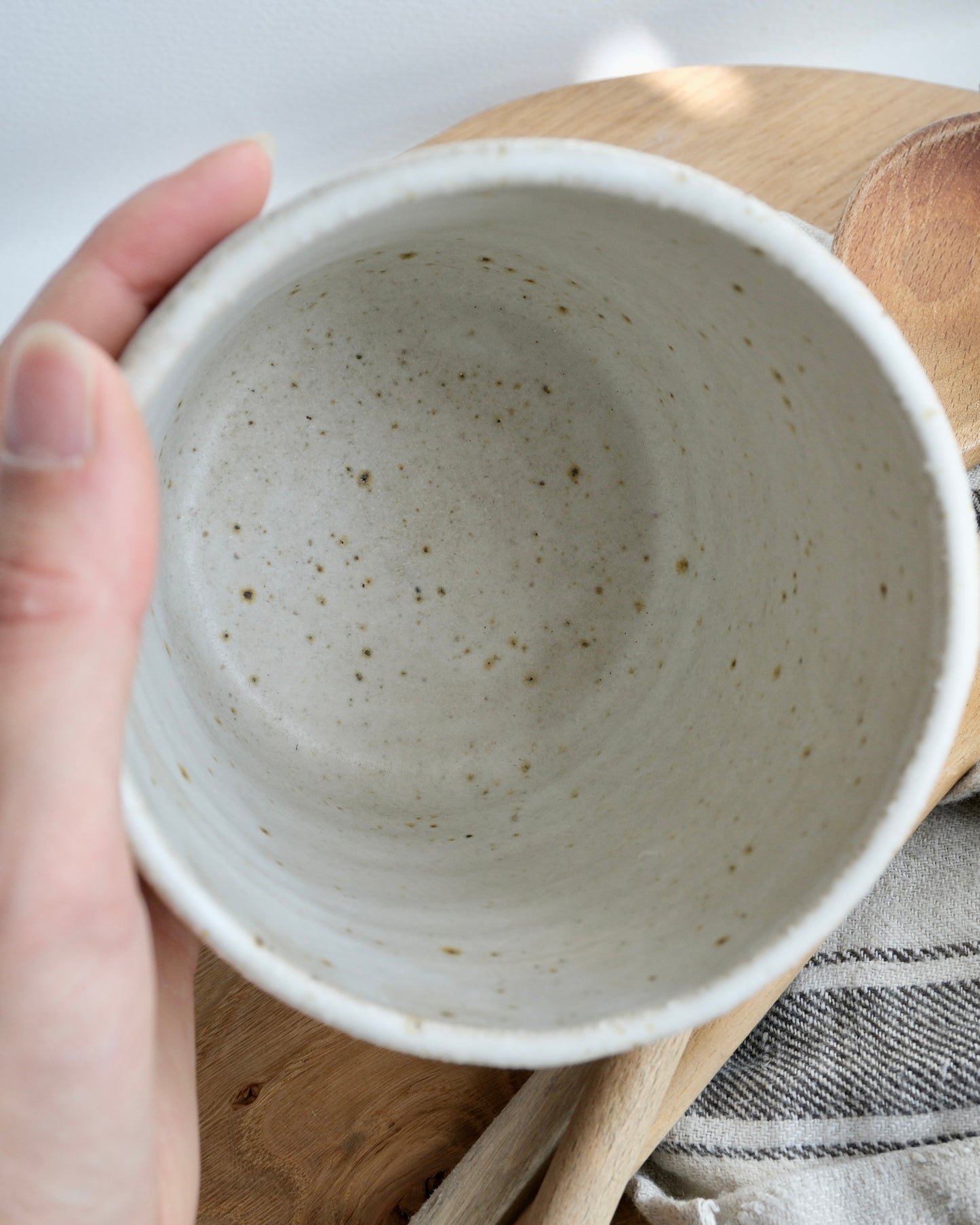 Hand holding a medium ceramic pot with a speckled texture on a wooden surface.
