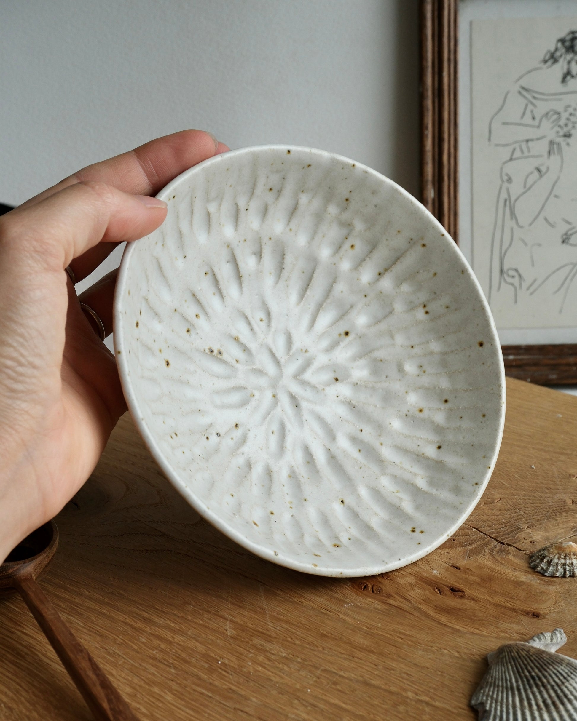Handmade white ceramic dish bowl with a carved design on a wooden surface with a framed picture in the background