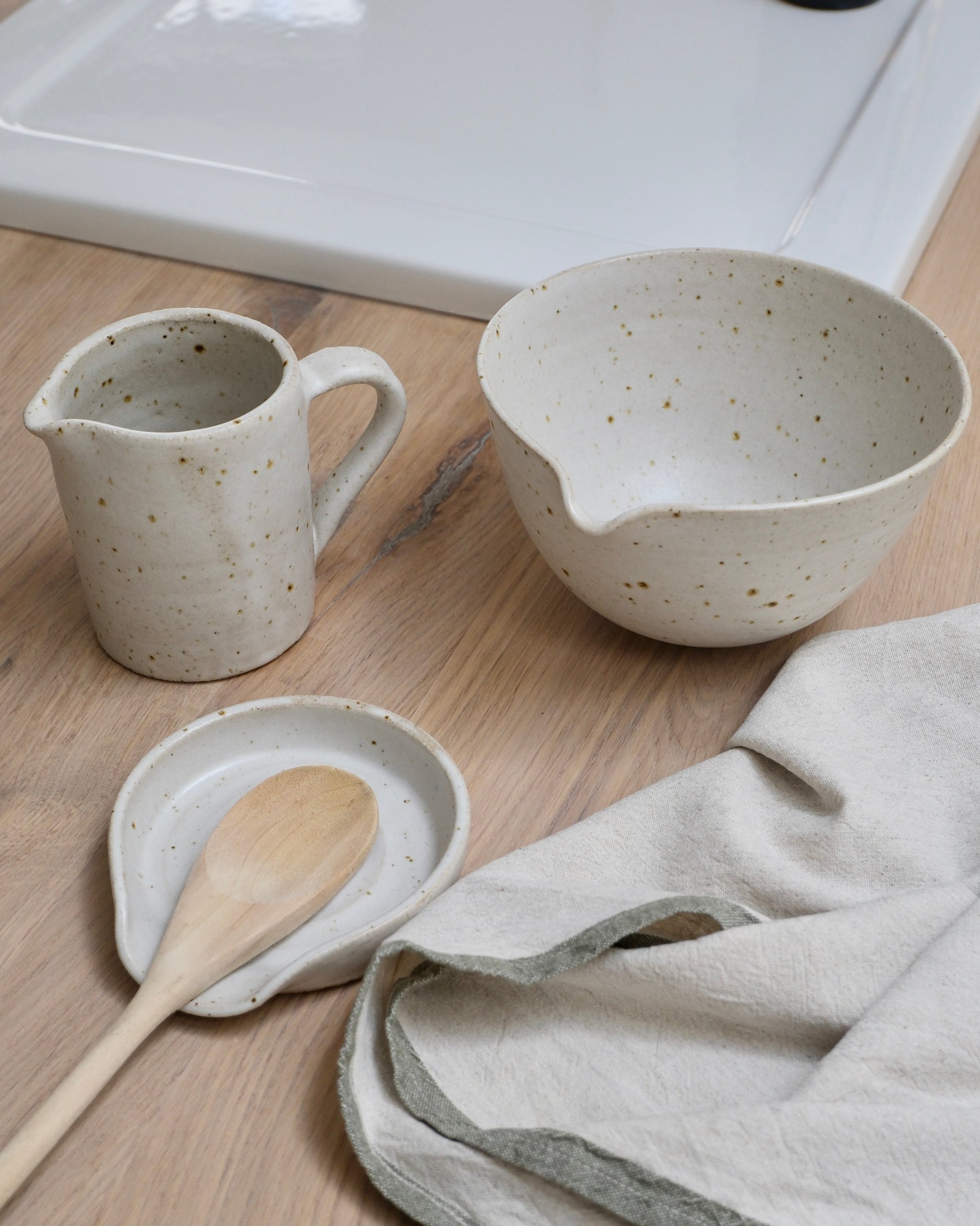Set of white speckled ceramic kitchenware including a pitcher, mixing bowl, and small plate with a wooden spoon on a wooden surface.