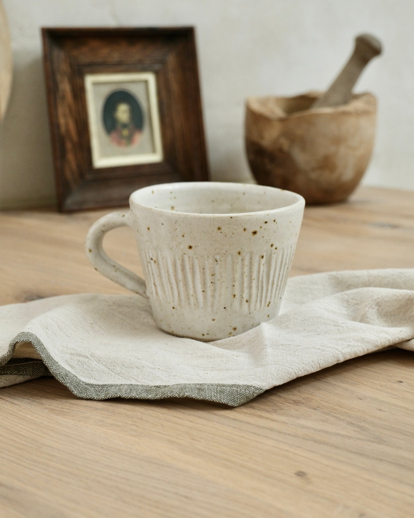 white Ceramic coffee mug with textured line striped design on a wooden surface with a cloth, framed picture, and wooden bowl in the background.