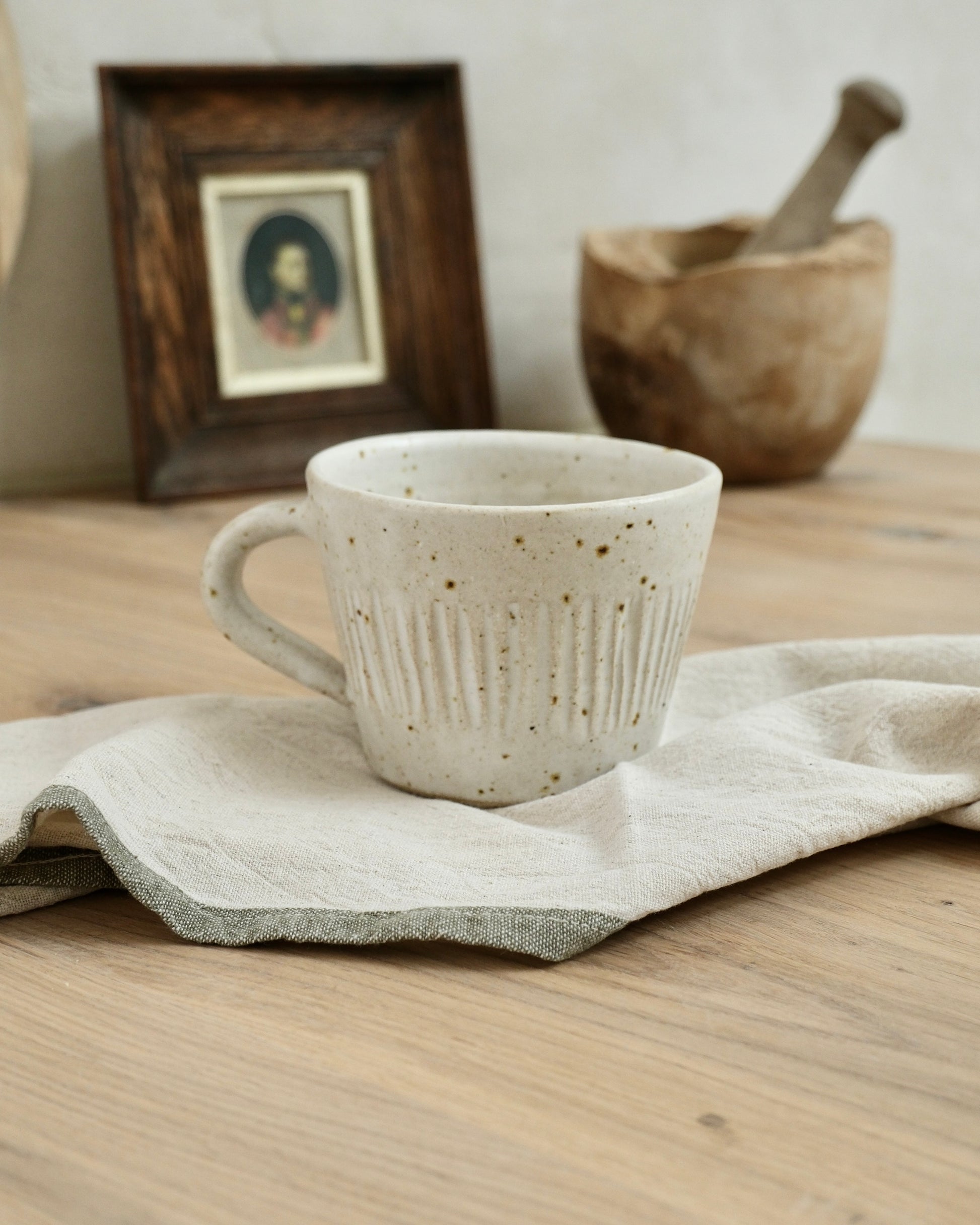 white Ceramic coffee mug with textured line striped design on a wooden surface with a cloth, framed picture, and wooden bowl in the background.