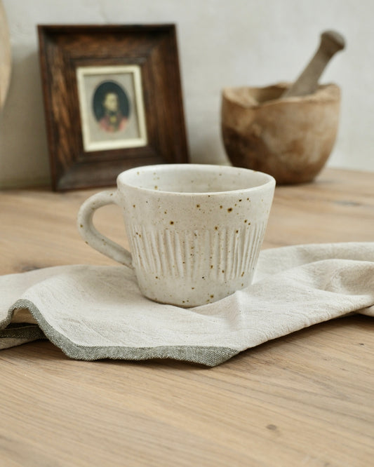 white Ceramic coffee mug with textured line striped design on a wooden surface with a cloth, framed picture, and wooden bowl in the background.