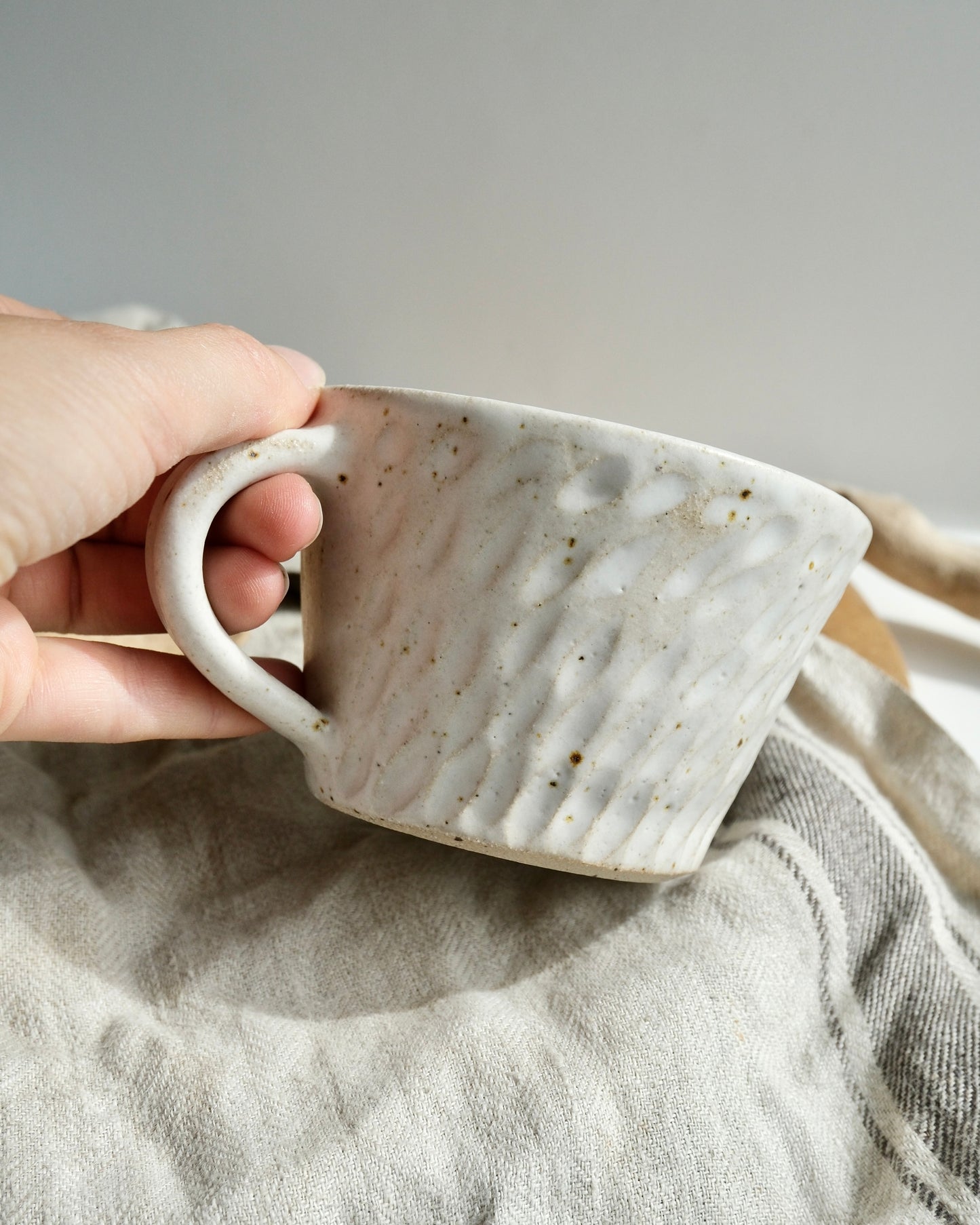 Hand holding a white textured ceramic coffee mug with a carved pattern on a tea towel
