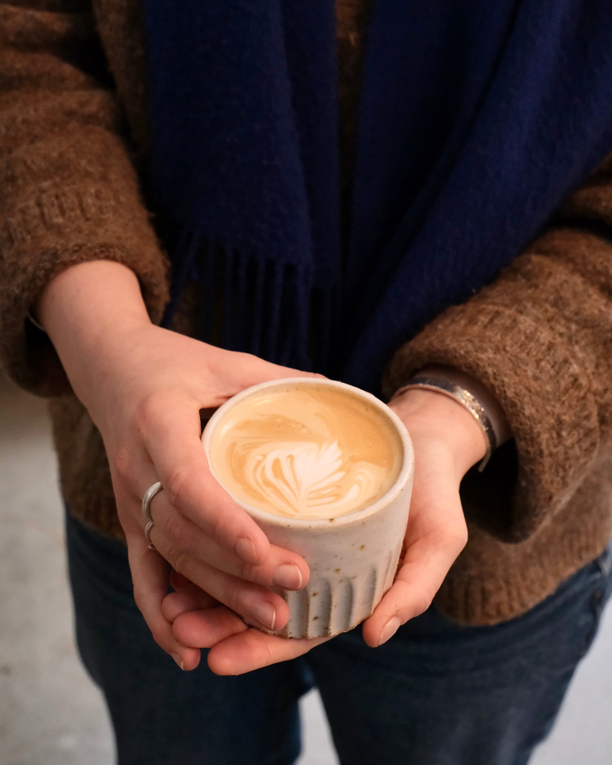 Person holding a handmade white ceramic cup of coffee with latte art, wearing a brown sweater and dark blue scarf.