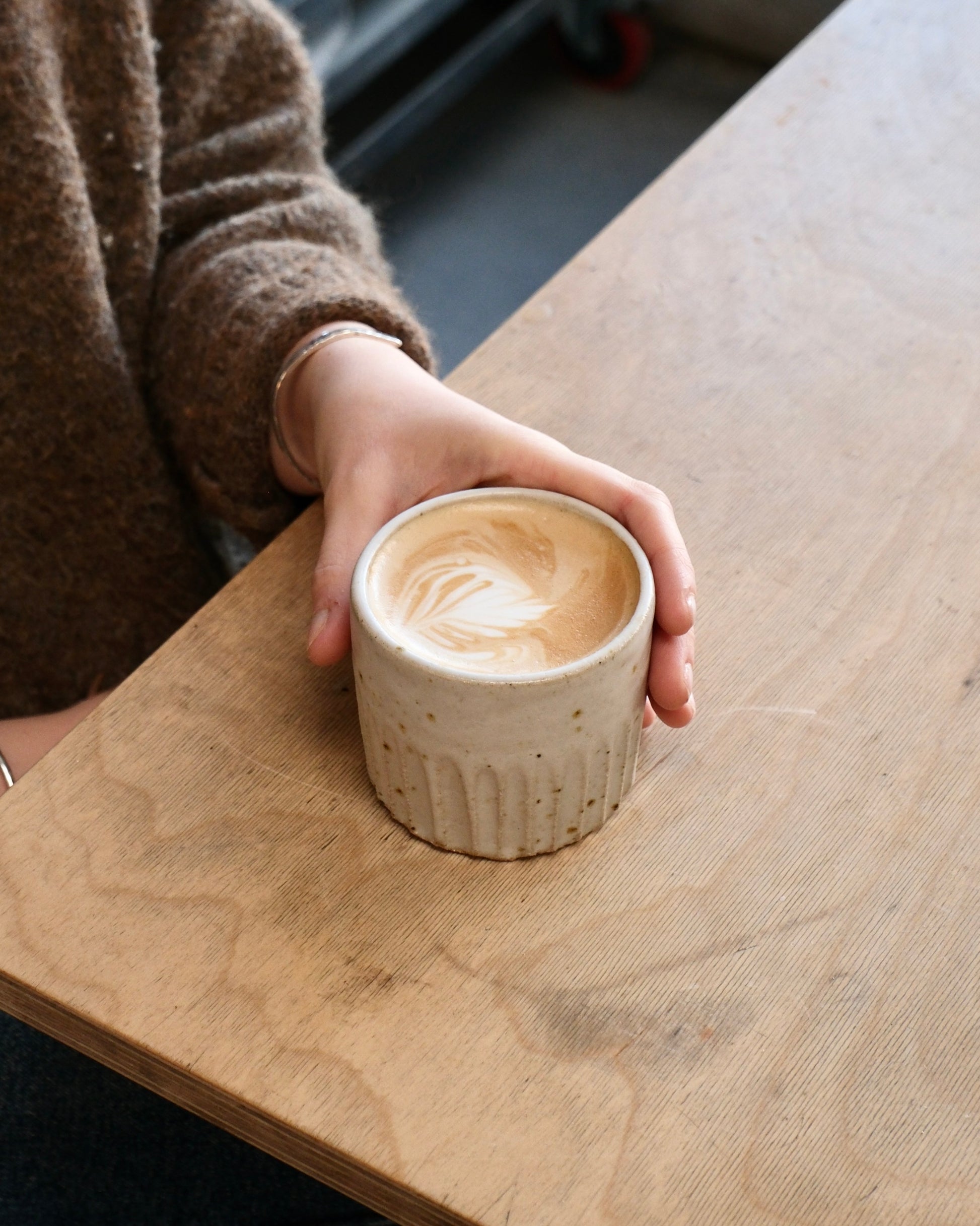 Person holding a white ceramic cup with a latte art design on a wooden table