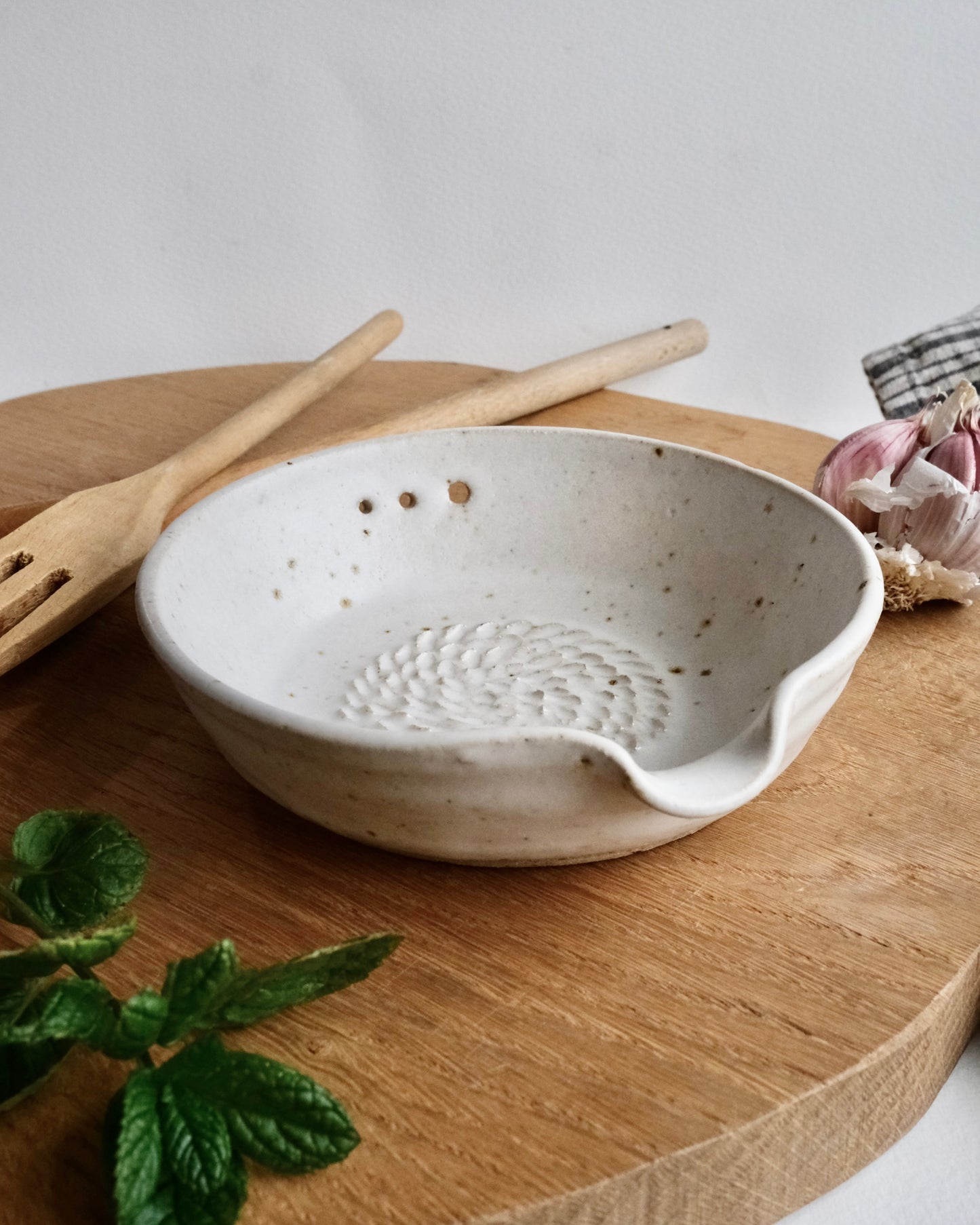 White ceramic food grater bowl bowl with textured design on a wooden surface, accompanied by a wooden spatula and a plant.