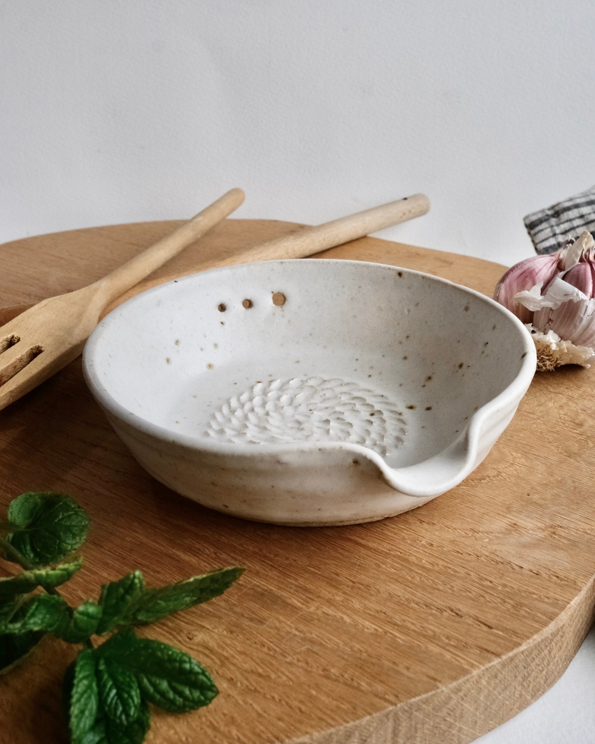 White ceramic food grater bowl bowl with textured design on a wooden surface, accompanied by a wooden spatula and a plant.