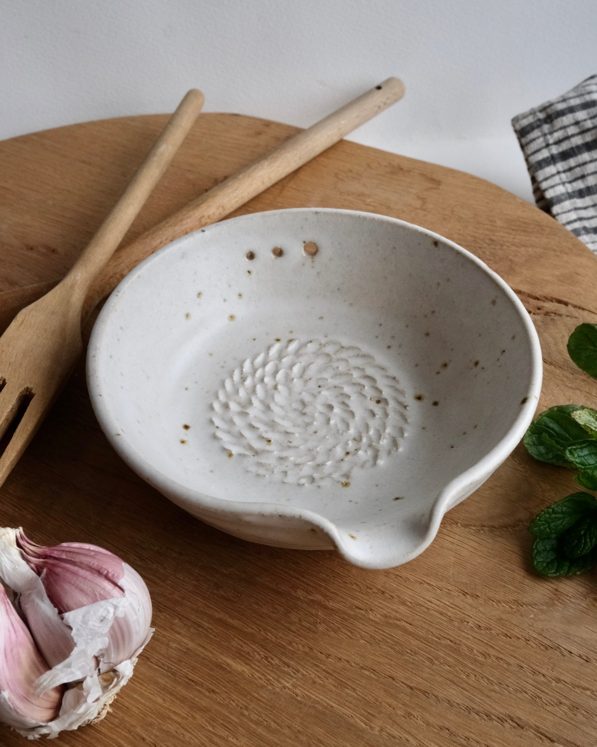 White ceramic garlic dish and herb striper on a wooden surface with a wooden spoon and garlic bulb.