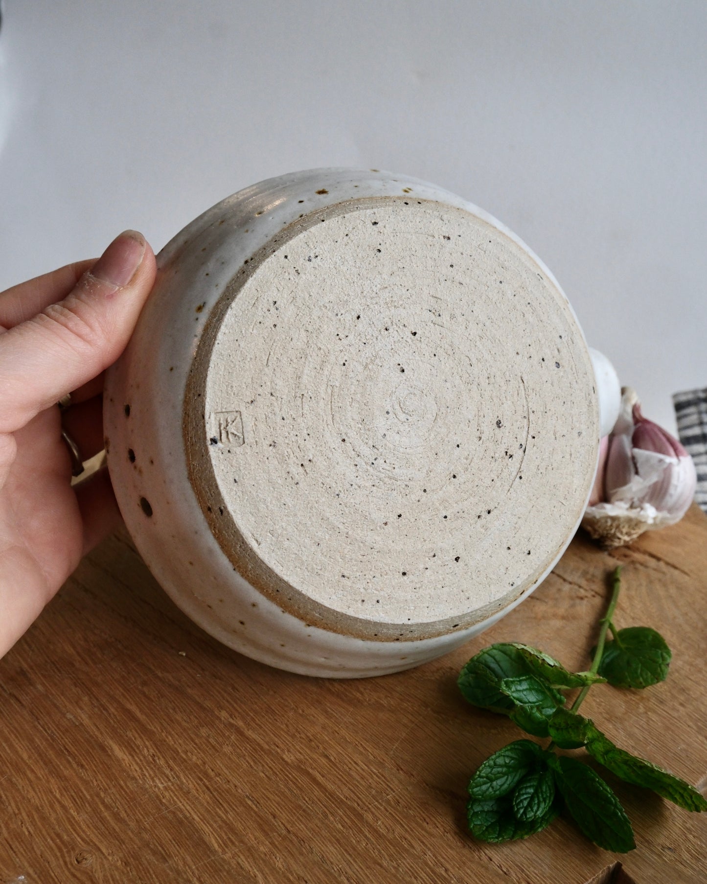 Ceramic pot with speckled glaze held by a hand on a wooden surface with a plant.