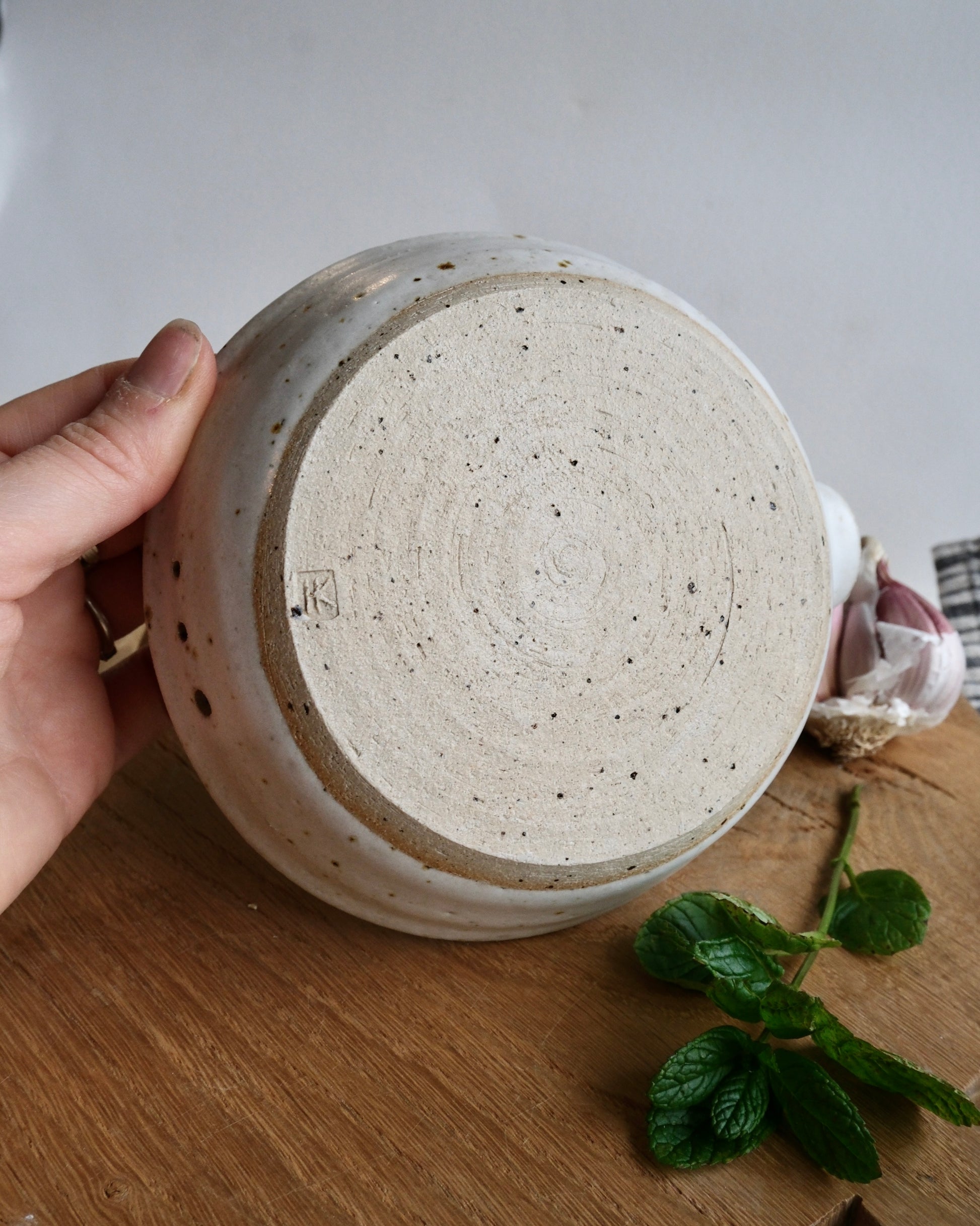 Ceramic pot with speckled glaze held by a hand on a wooden surface with a plant.
