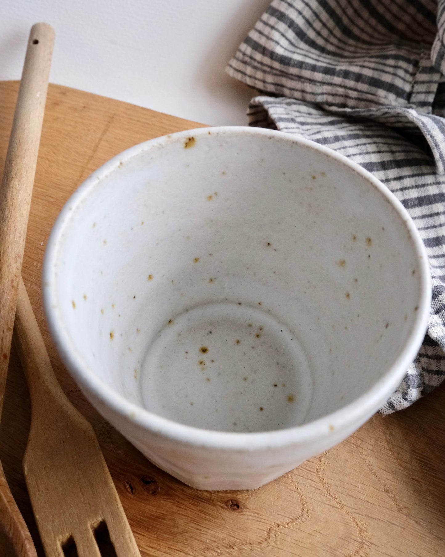 handmade White ceramic cup with speckled texture on a wooden surface with a striped cloth and wooden utensils.