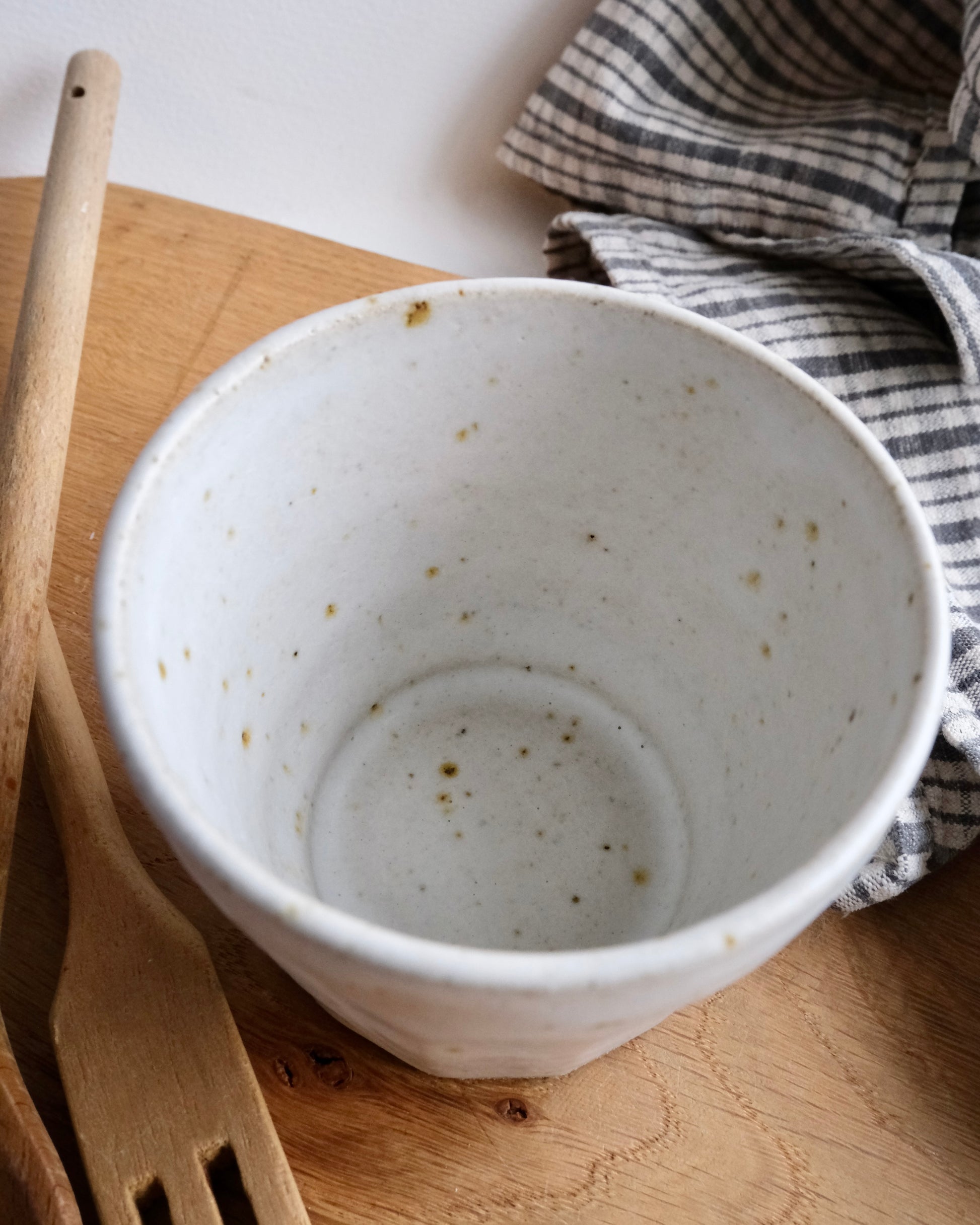 handmade White ceramic cup with speckled texture on a wooden surface with a striped cloth and wooden utensils.