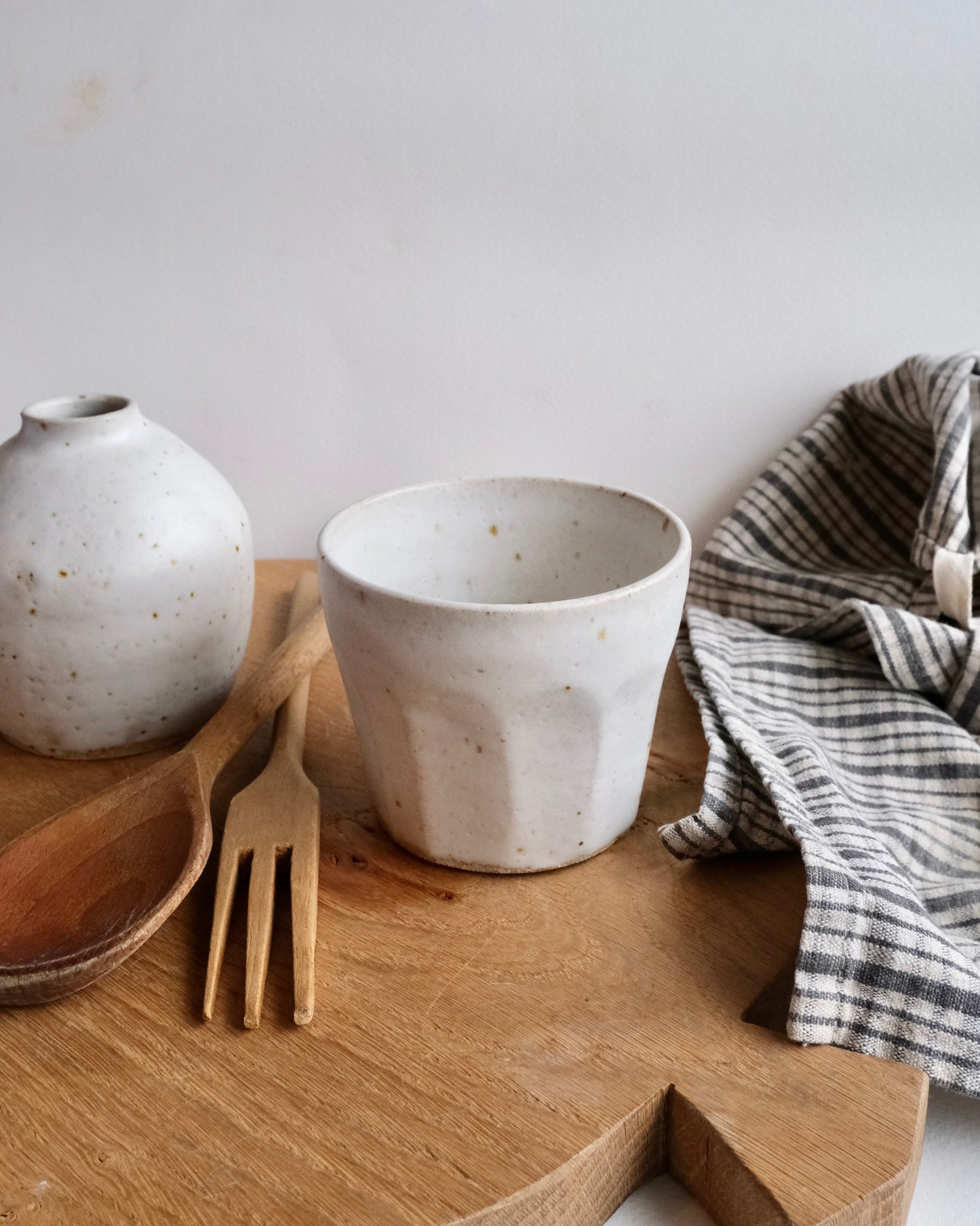 Ceramic cup and white bud vase, wooden spoon, and fork on a wooden surface with a white background